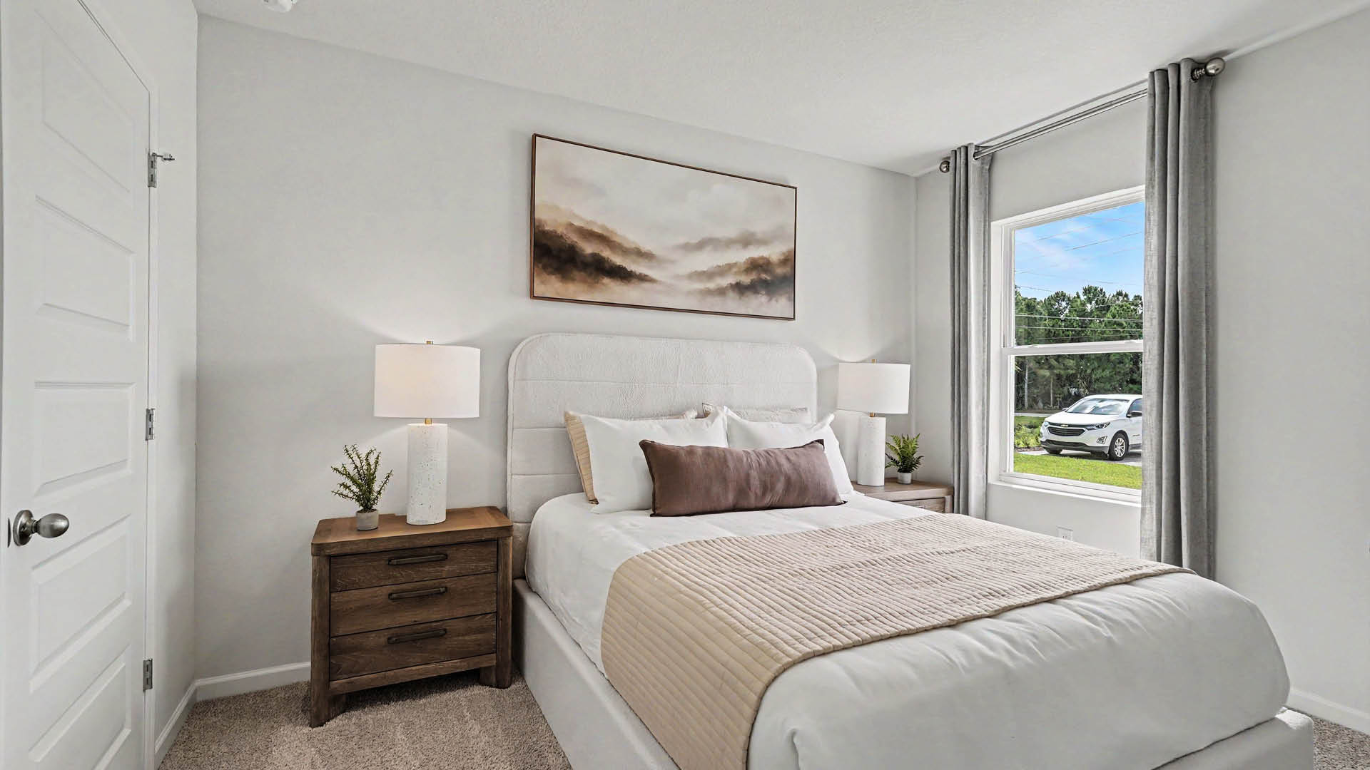 A cozy room with beige carpet, white walls, and a window, allowing natural light to illuminate the space. A closed door is visible.