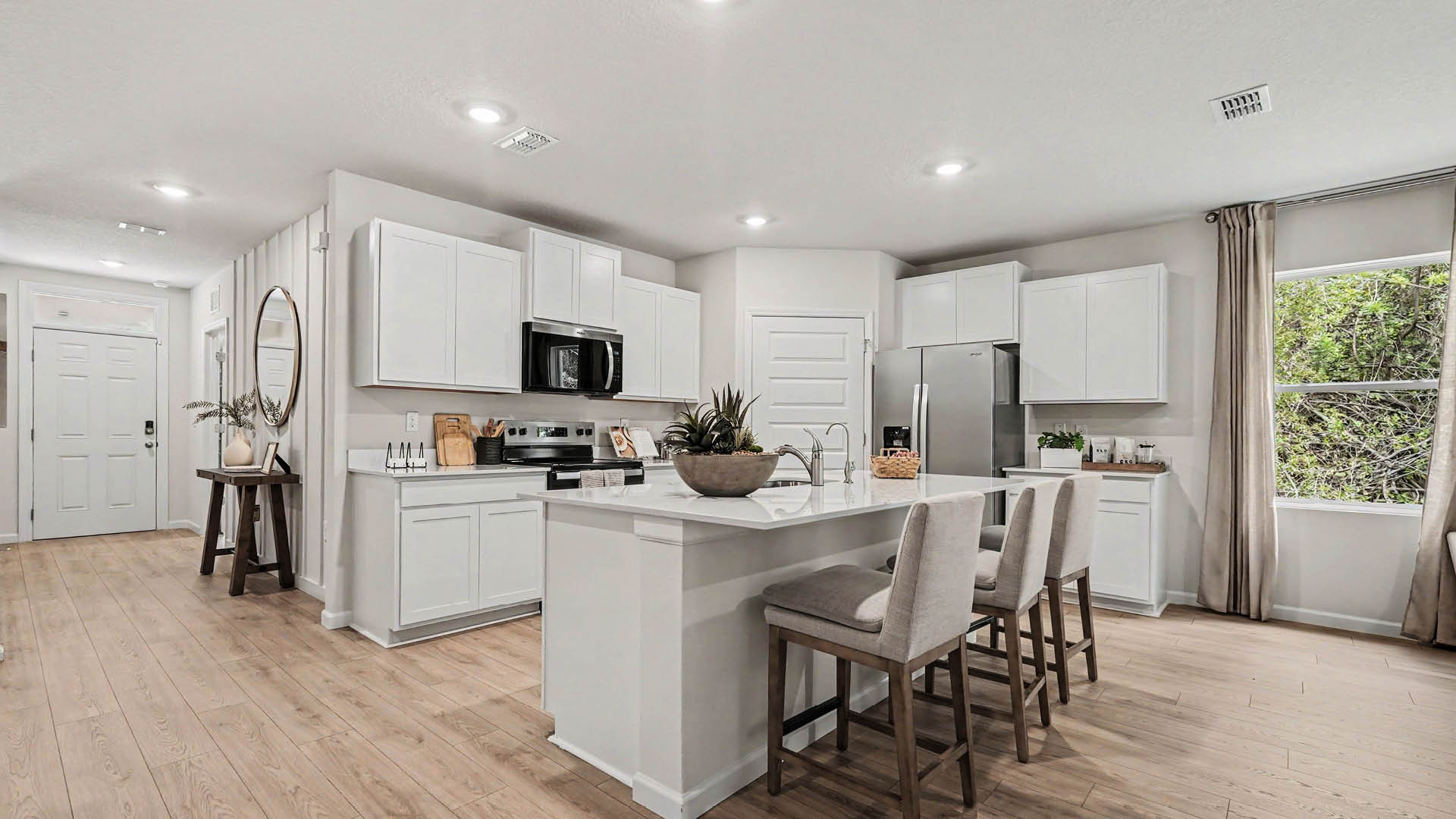 A large kitchen with stainless steel appliances and an island with seating. An entryway hallway leading to the front door is visible.