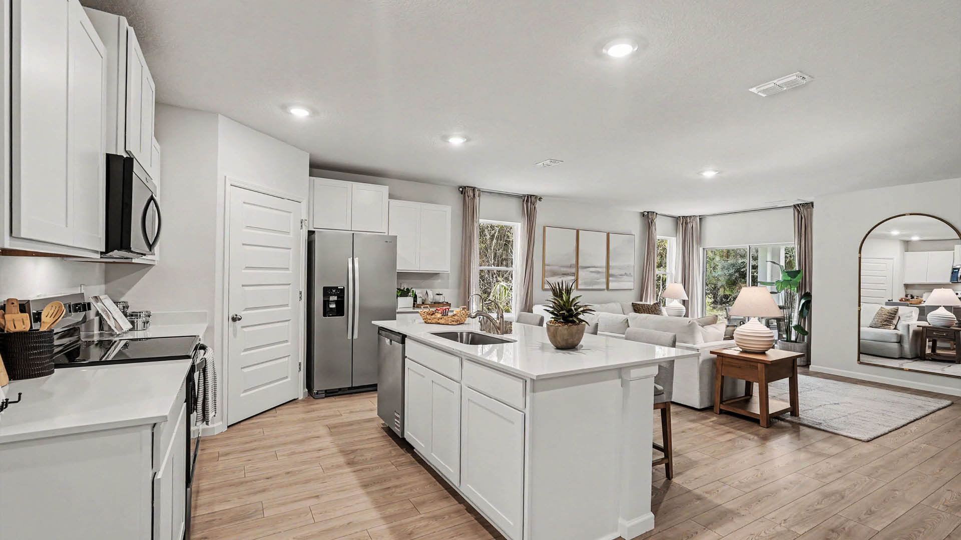A large kitchen with stainless steel appliances and an island with seating. A living room area featuring large windows and a sliding glass door are visible.