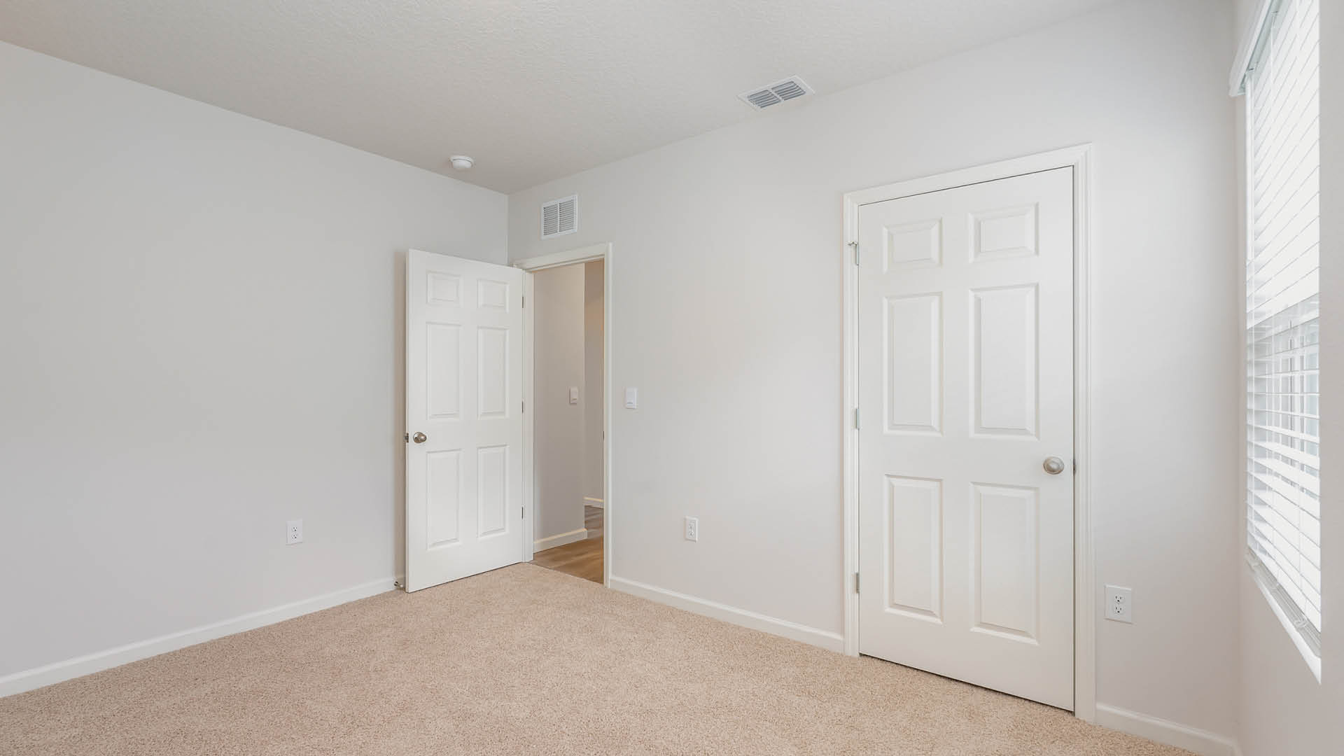 An empty room with beige carpet, white walls, and a window with blinds, allowing natural light to illuminate the space. The door to the room is open, while another door, leading to the closet, is closed.