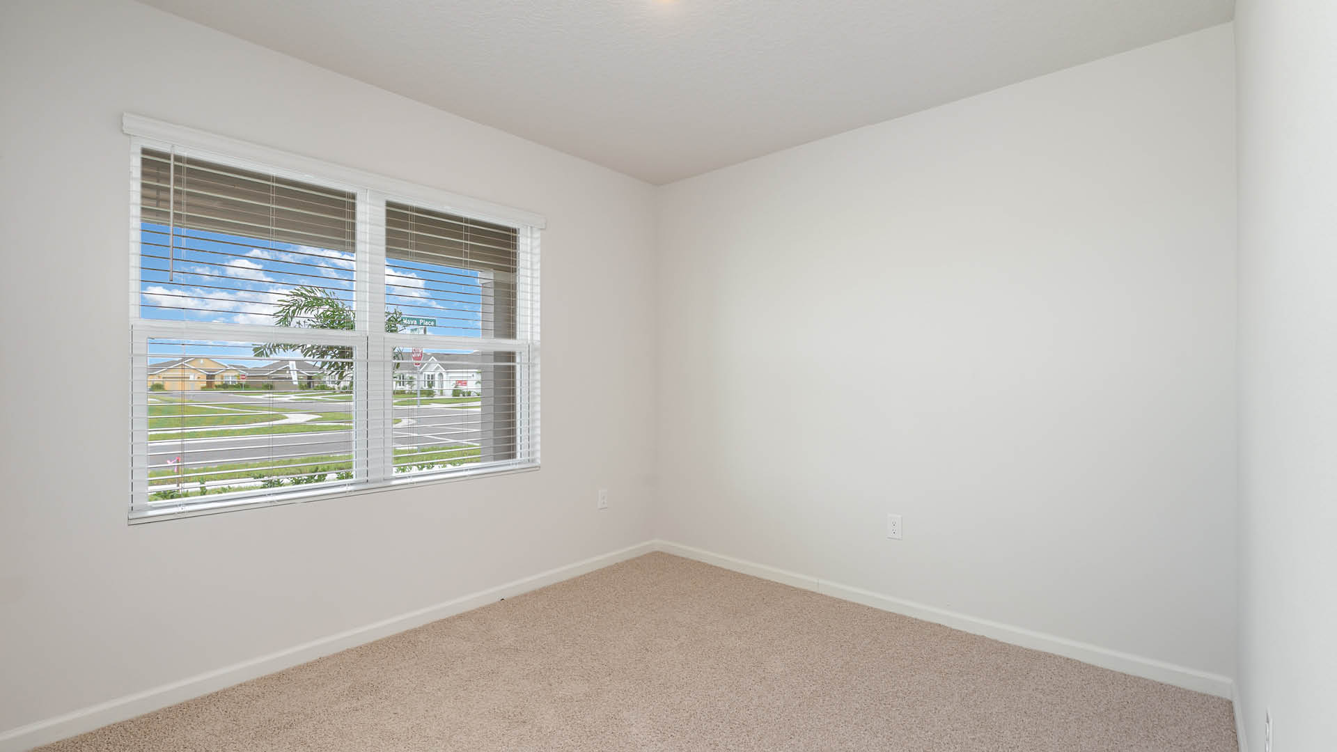 An empty room with beige carpet, white walls, and a large window with blinds, allowing natural light to illuminate the space.