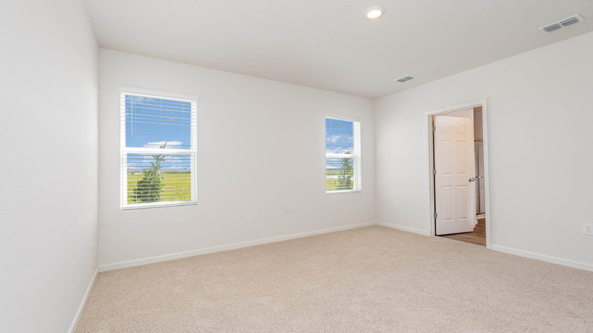 A large bedroom with beige carpet, white walls, and two windows, allowing natural light to illuminate the space. An open door leading to an ensuite bathroom is visible.