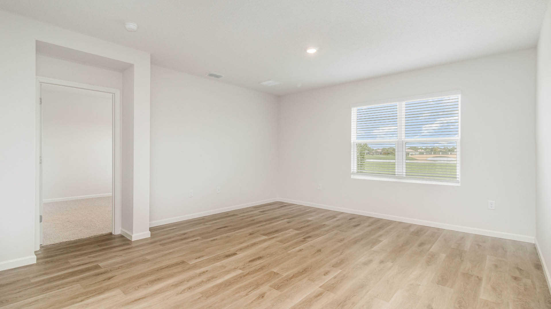 A spacious, empty living room with a large window and minimalistic walls. An open door leading to a bedroom is visible.
