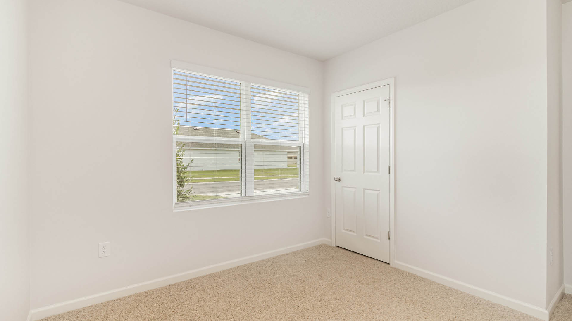 An empty room with beige carpet, white walls, and a window with blinds, allowing natural light to illuminate the space. A closed door is visible.