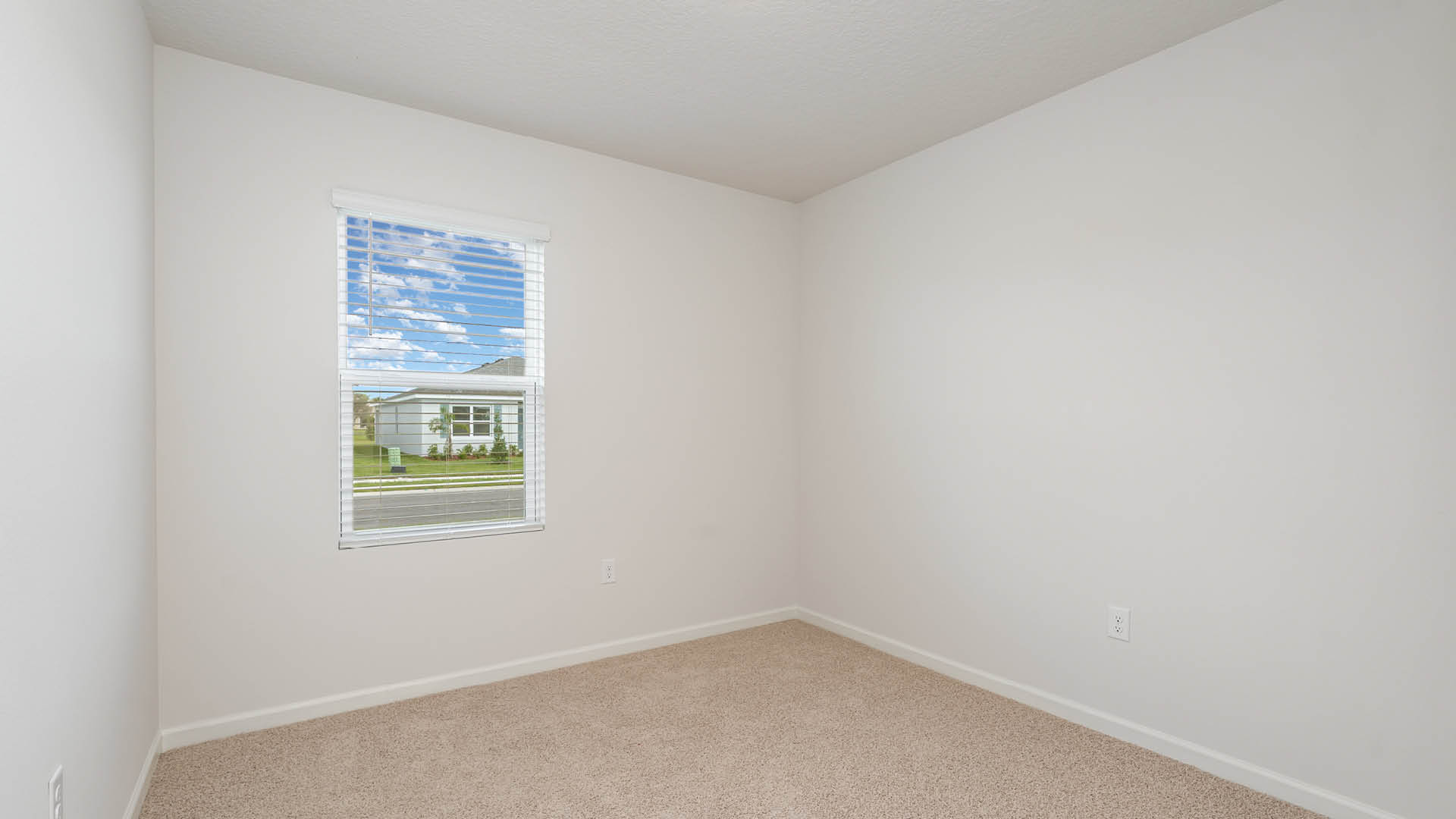 An empty room with beige carpet, white walls, and a window with blinds, allowing natural light to illuminate the space.