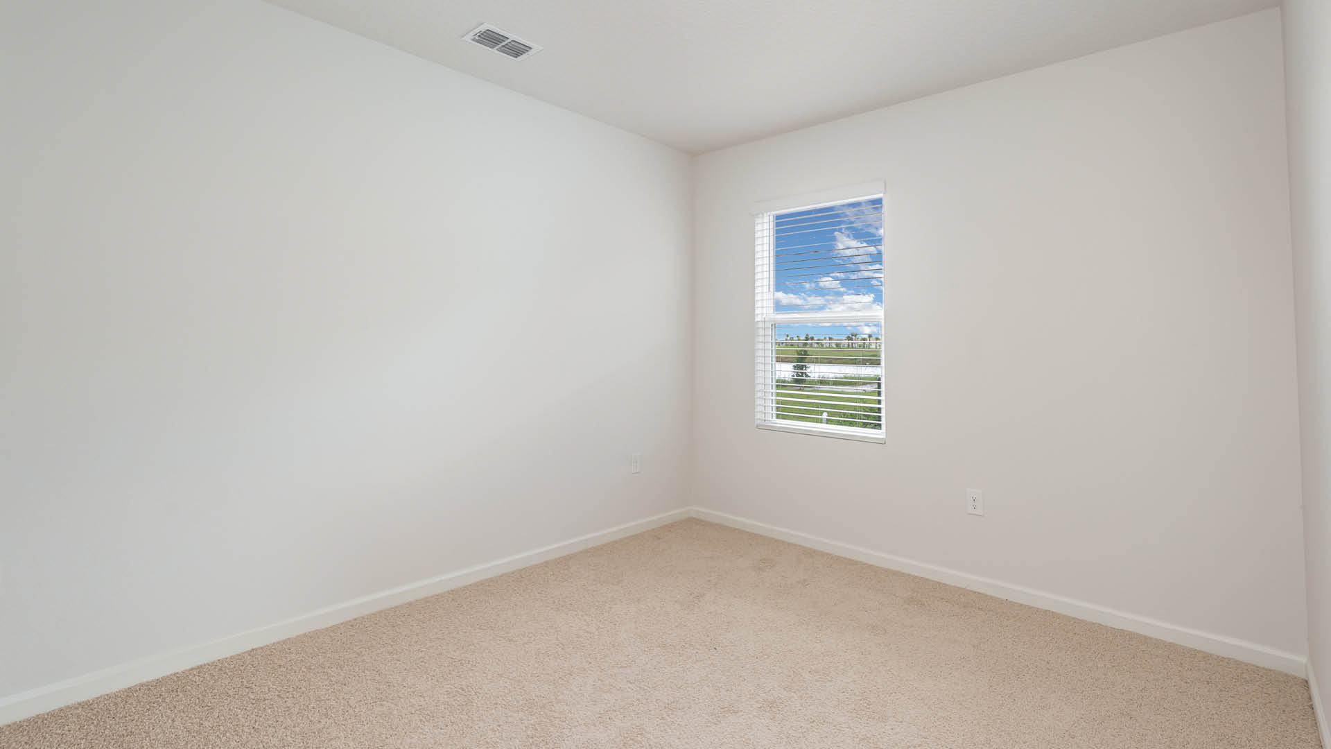 An empty room with beige carpet, white walls, and a window with blinds, allowing natural light to illuminate the space.