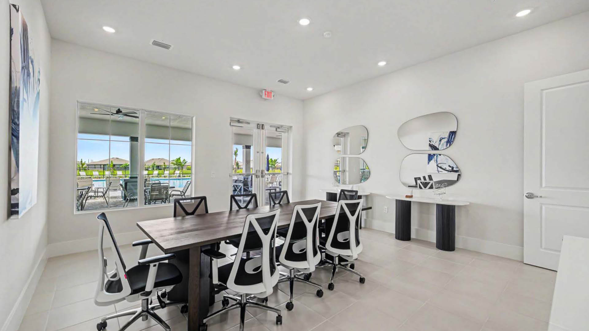 A collaboration space with a large table and chairs. Windows and a glass door show the pool and deck area outside.