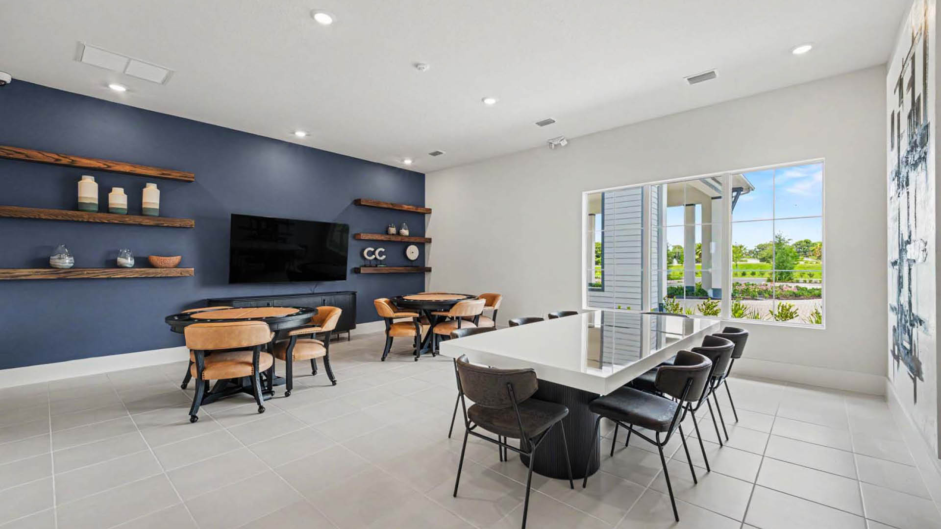 A gathering space with tables and chairs to accommodate different size groups. A TV is mounted to the wall and the windows look out onto a beautifully landscaped area.