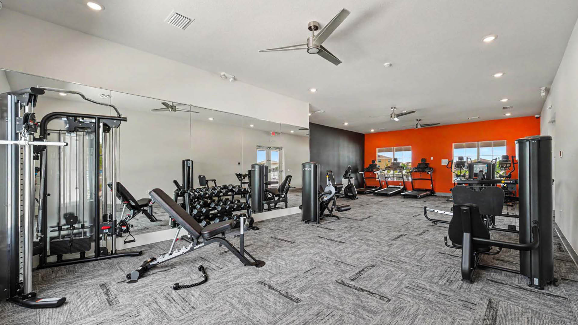 A fitness center with ceiling fans and a mirrored wall. Large windows let in natural light.