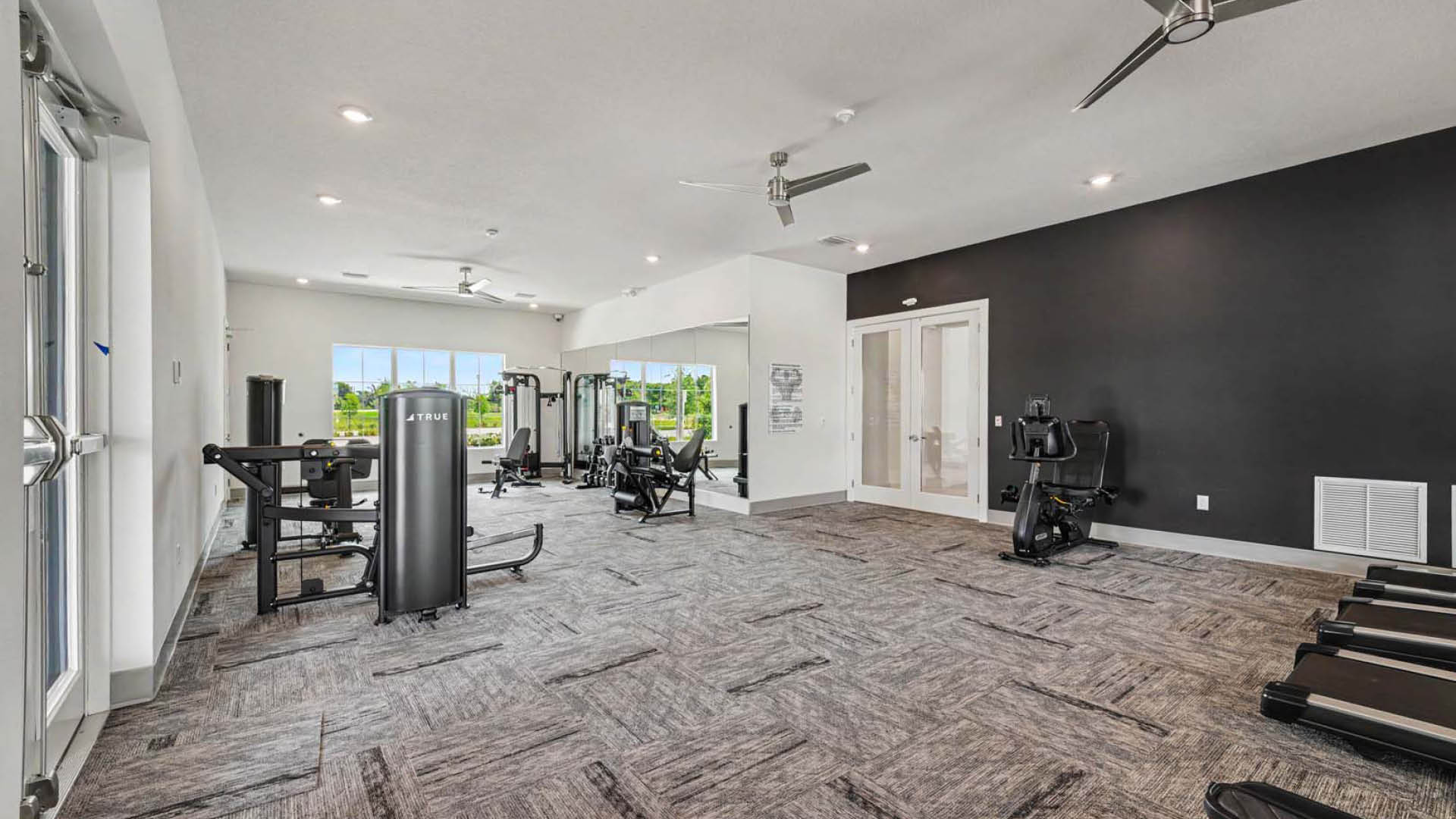 A fitness center with ceiling fans and a mirrored wall. Large windows let in natural light.