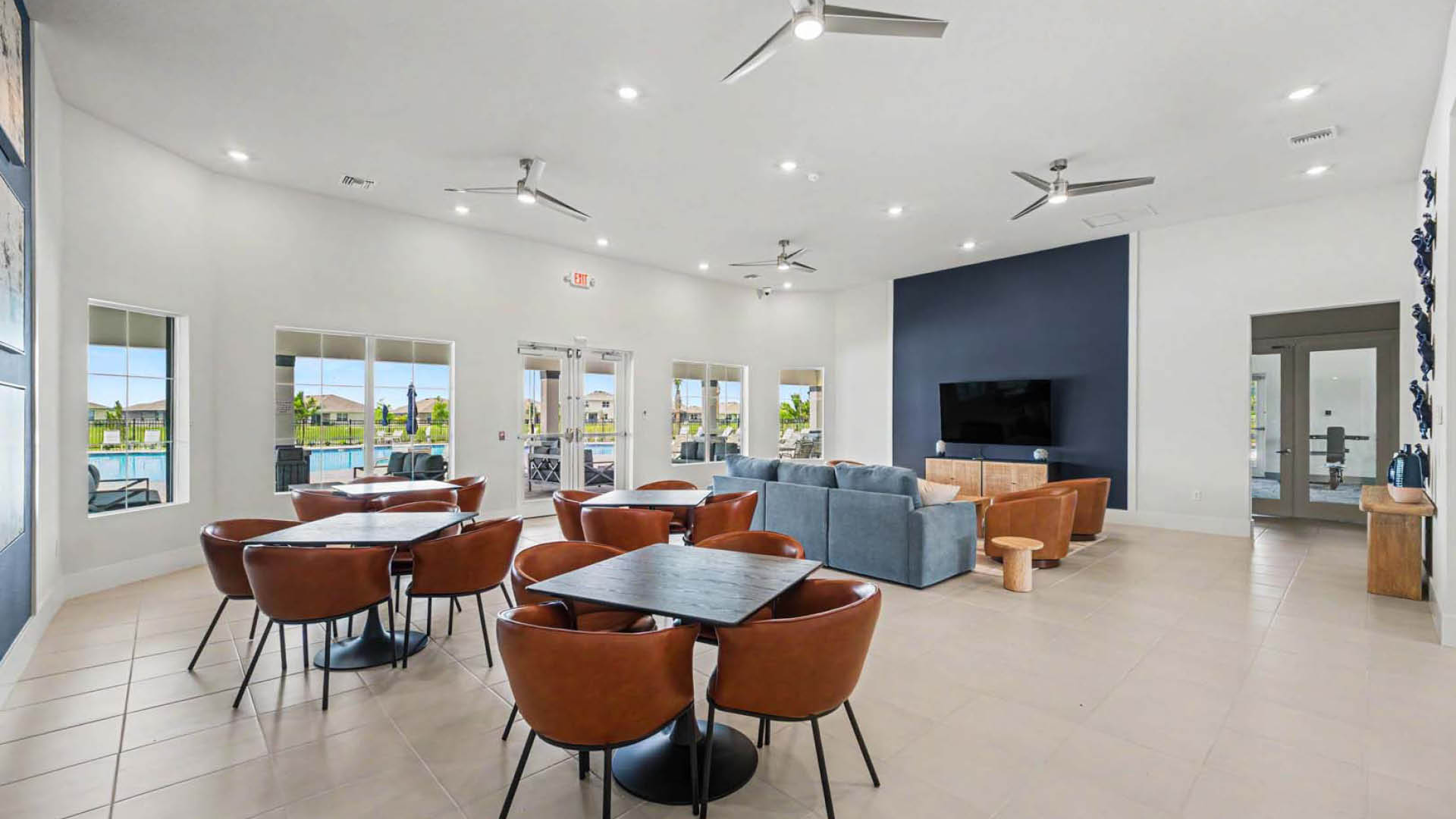 A large gathering area with tables and chairs, couches, lounge areas and a TV. Several large windows and glass doors lookout onto a pool and deck area. A room with fitness equipment is visible.