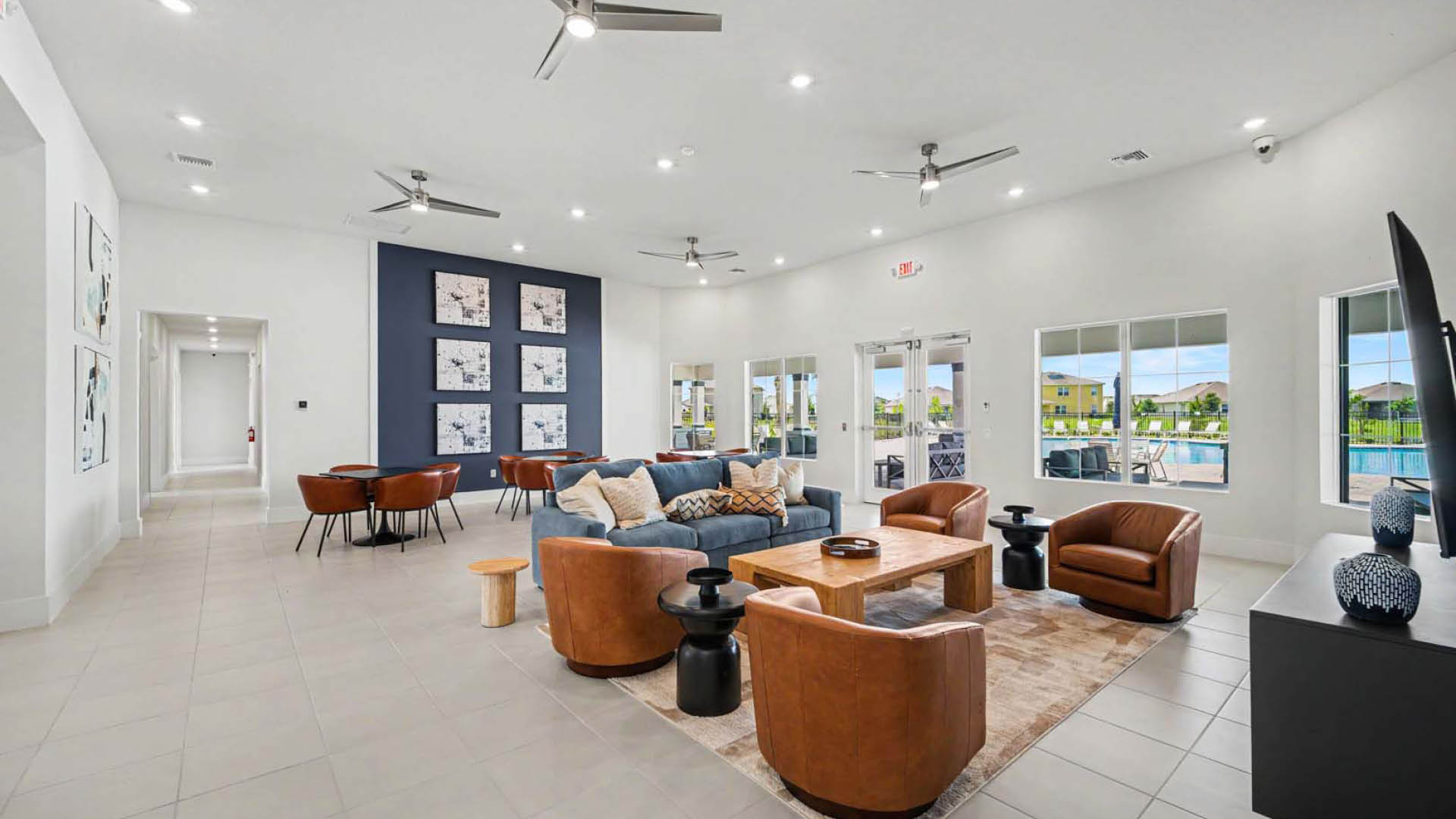 A large gathering area with tables and chairs, couches, lounge areas and a TV. Several large windows and glass doors lookout onto a pool and deck area.