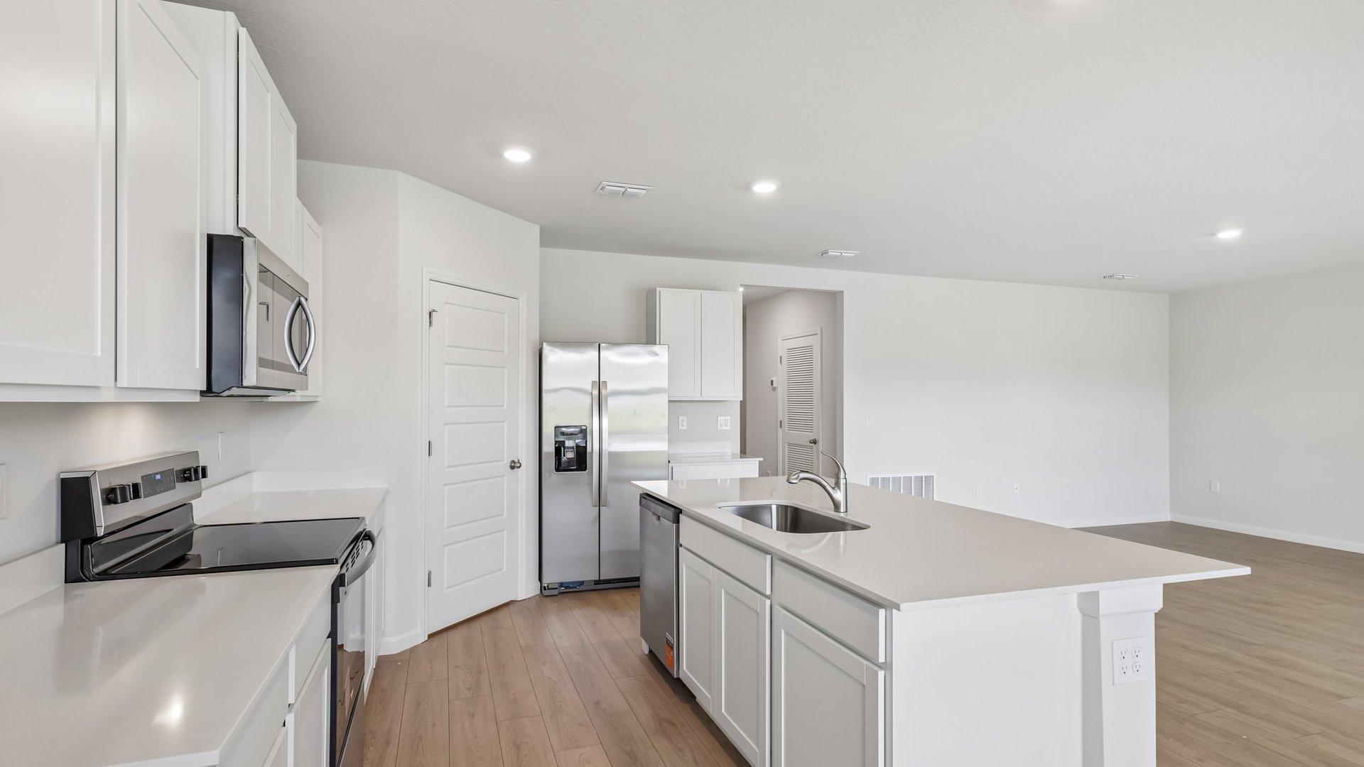 A modern kitchen with white cabinetry, stainless steel appliances, and a large island. A large living area and an opening to another area are visible.
