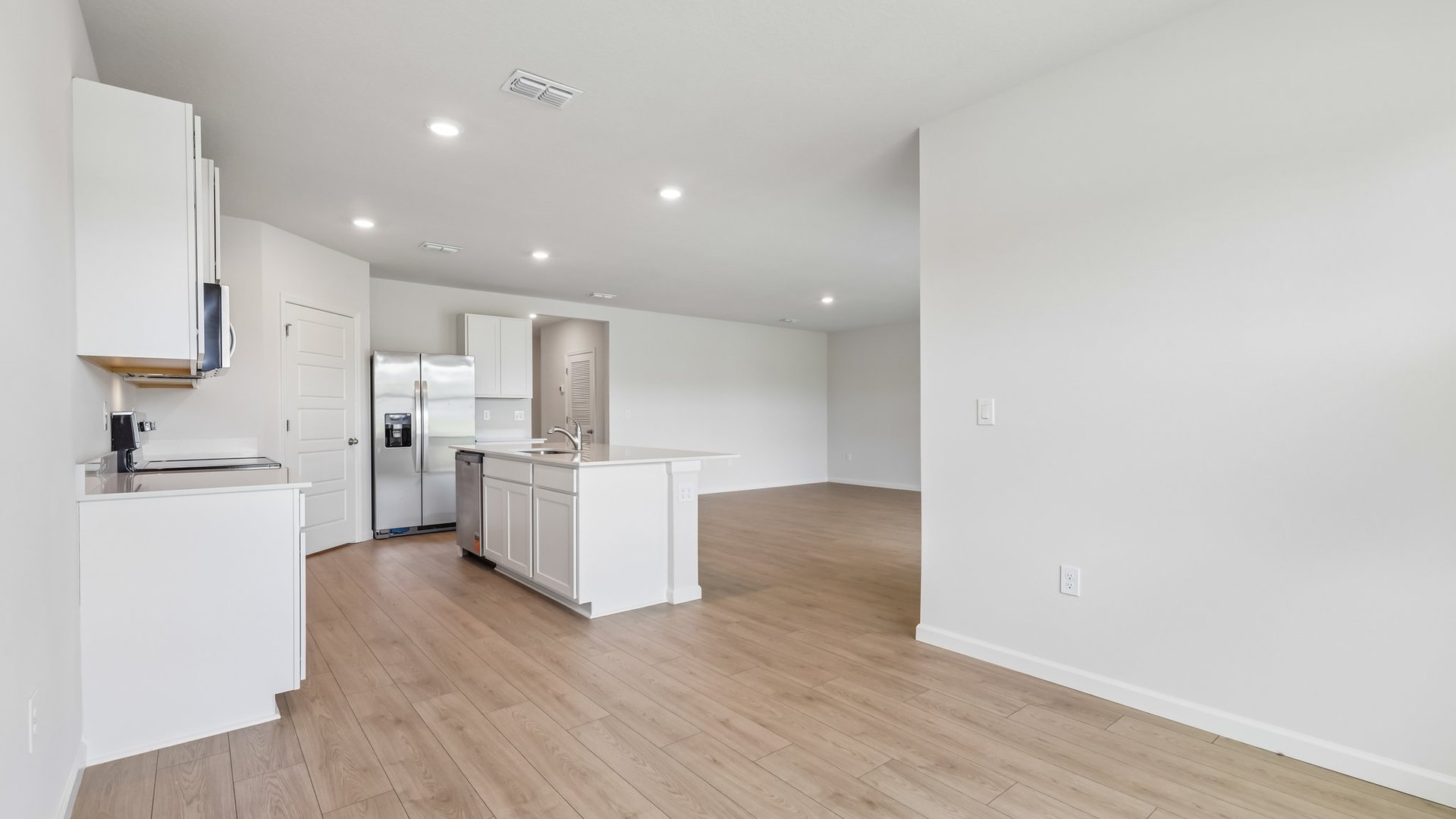The view of the open concept home from the dining area. The kitchen is visible with quartz countertops, stainless steel appliances, and an island with room for seating. A large living room area is also visible.