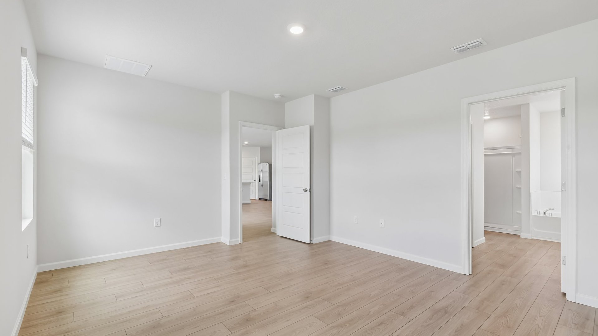 A large bedroom with windows, allowing natural light to illuminate the space. There is an open door to a large ensuite bathroom. The open door to the room is visible, showing the space beyond.