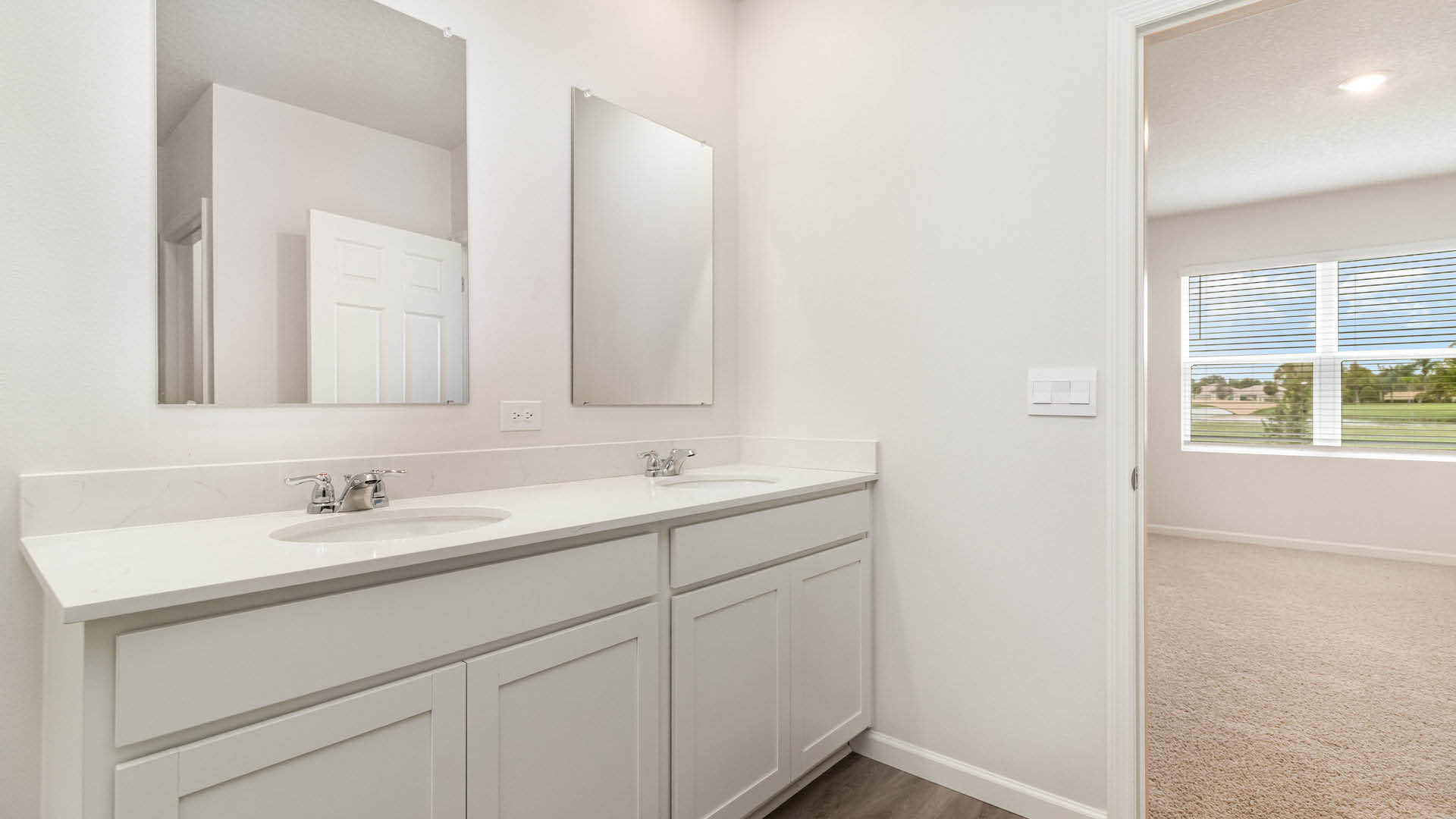 A spacious, ensuite bathroom featuring a double sink vanity with quartz countertops. The door to the bathroom is open, showing the attached bedroom.