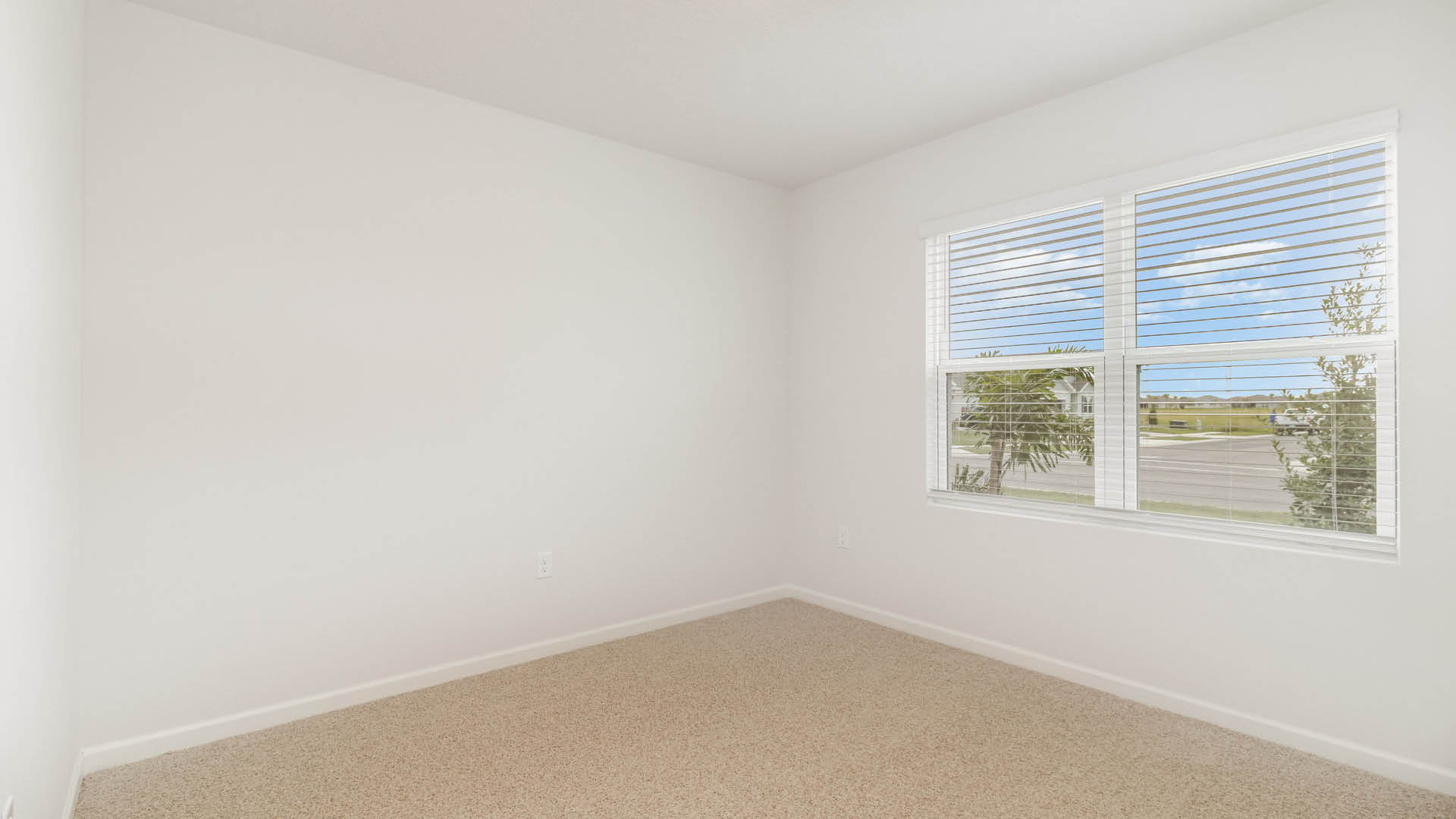 An empty room with beige carpet, white walls, and a large window with blinds, allowing natural light to illuminate the space.
