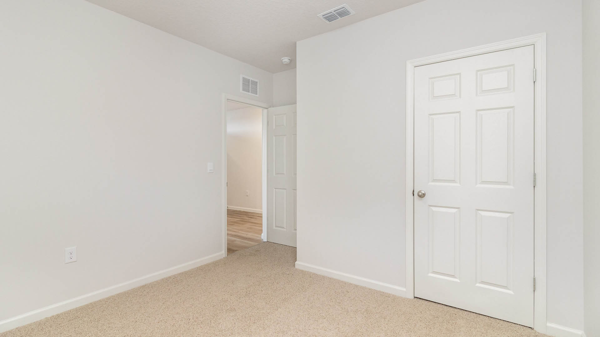 An empty room with beige carpet and white walls. The door to the room is open, while another door, leading to the closet, is closed.