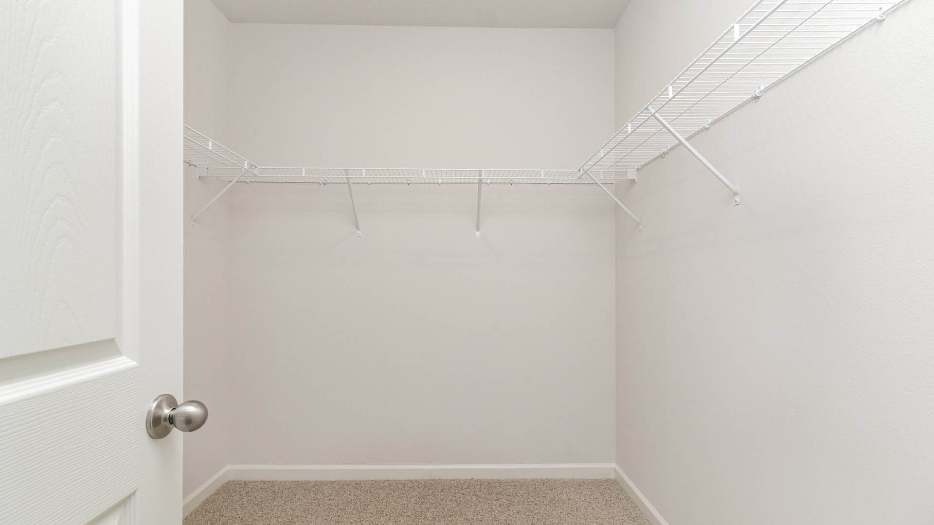 A spacious, walk-in closet with beige carpet, featuring white wire shelves against a light-colored wall.