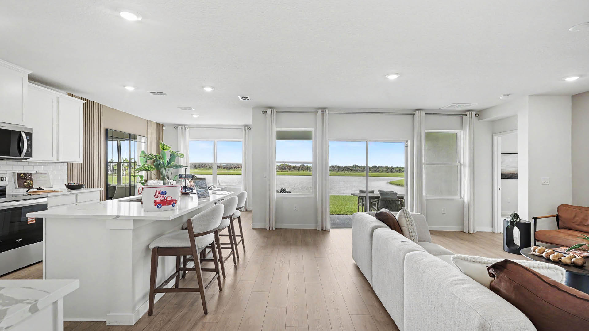 A modern open-concept living space features a white kitchen island with bar stools, a light-colored sectional sofa, and large windows and sliding glass door offering a view of a grassy outdoor area and water.