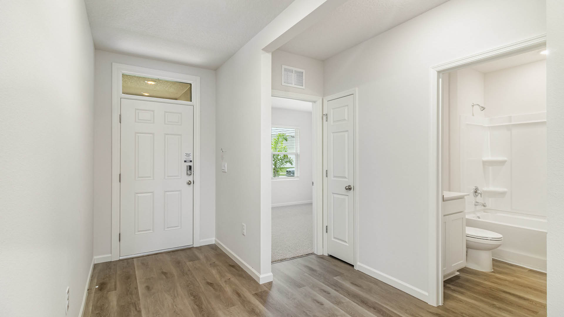 A white-walled entryway with light wood-look flooring features a front door on the left and openings leading to a carpeted bedroom, a closed closet, and a bathroom on the right.