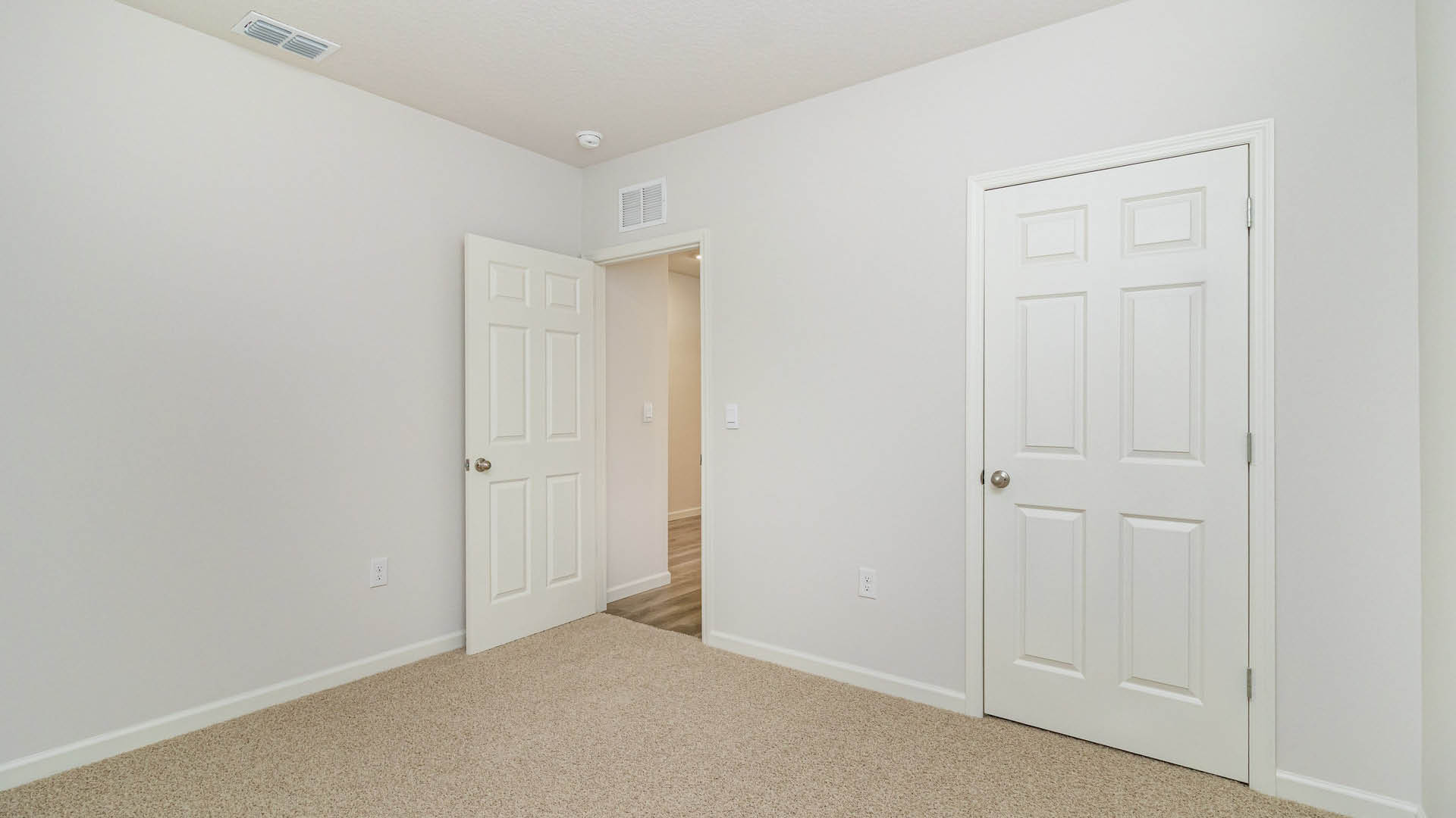A spacious, empty room with white walls and beige carpet, featuring a closed closet door and a hallway leading to another area.