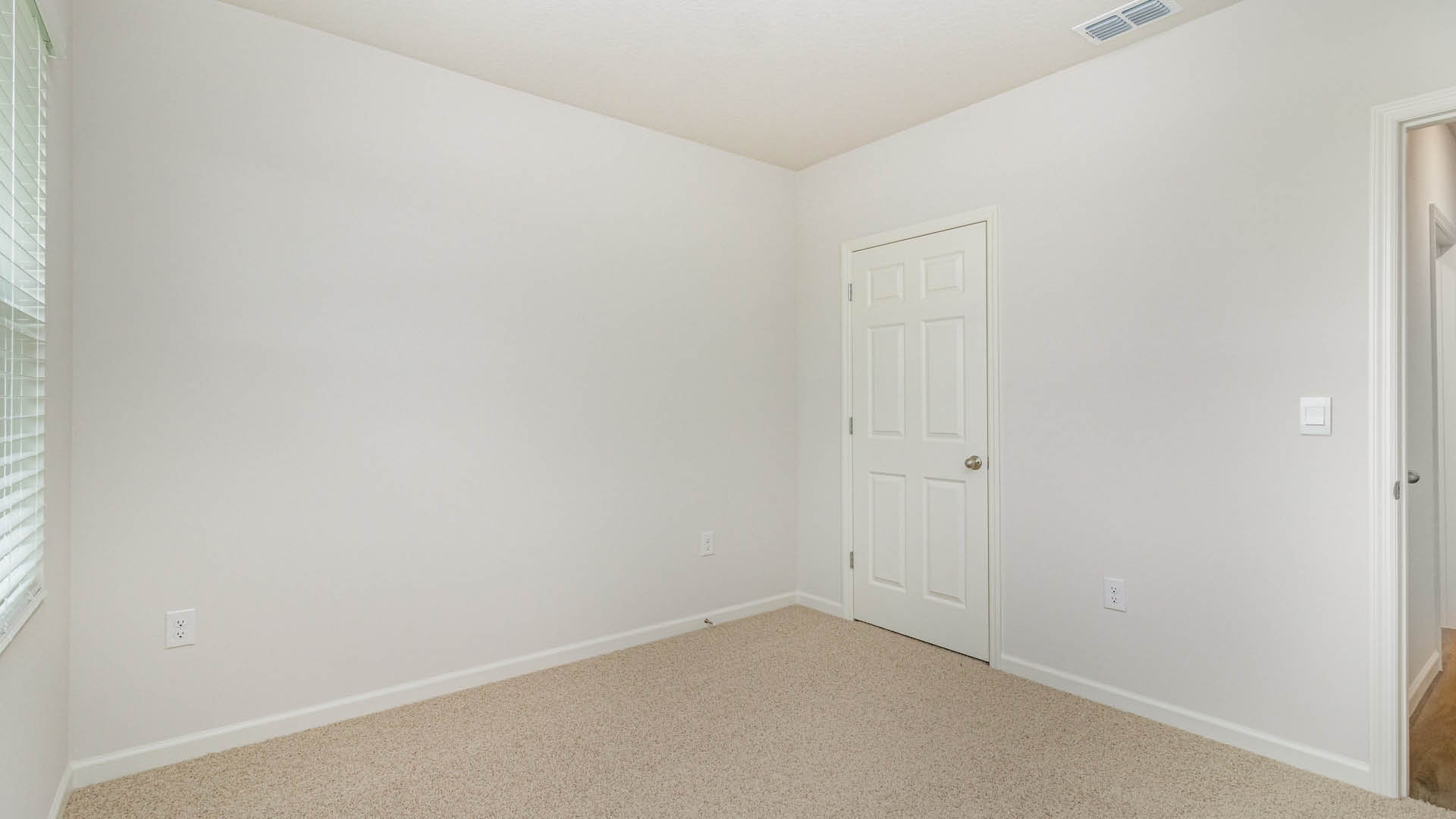 A spacious, empty room with white walls and beige carpet, featuring a closed closet door, an open door and hallway leading to another area, and a window with blinds.