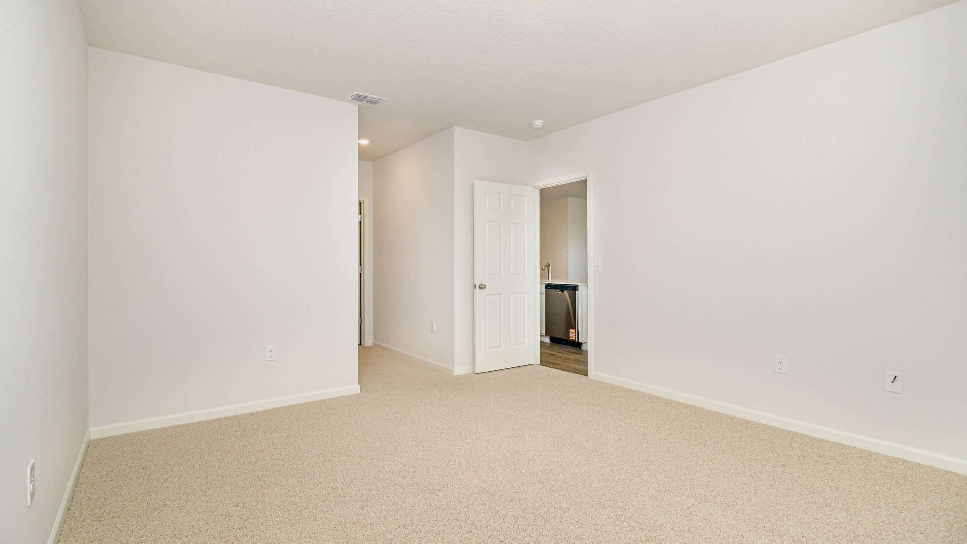 A spacious, empty room with white walls and beige carpet, featuring an open door and a hallway leading to another area.
