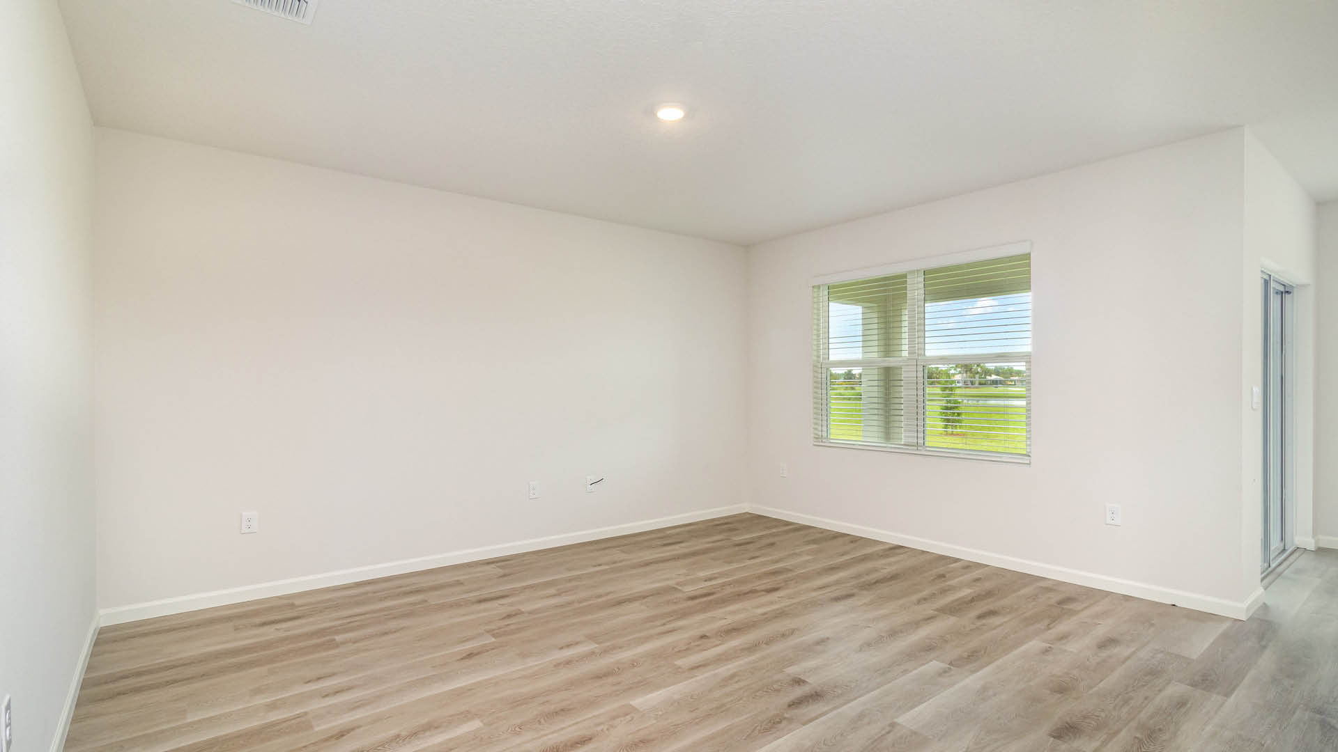 An empty, white-walled room with light brown LVP flooring features a single recessed ceiling light and a window with blinds looking out at a grassy landscape.