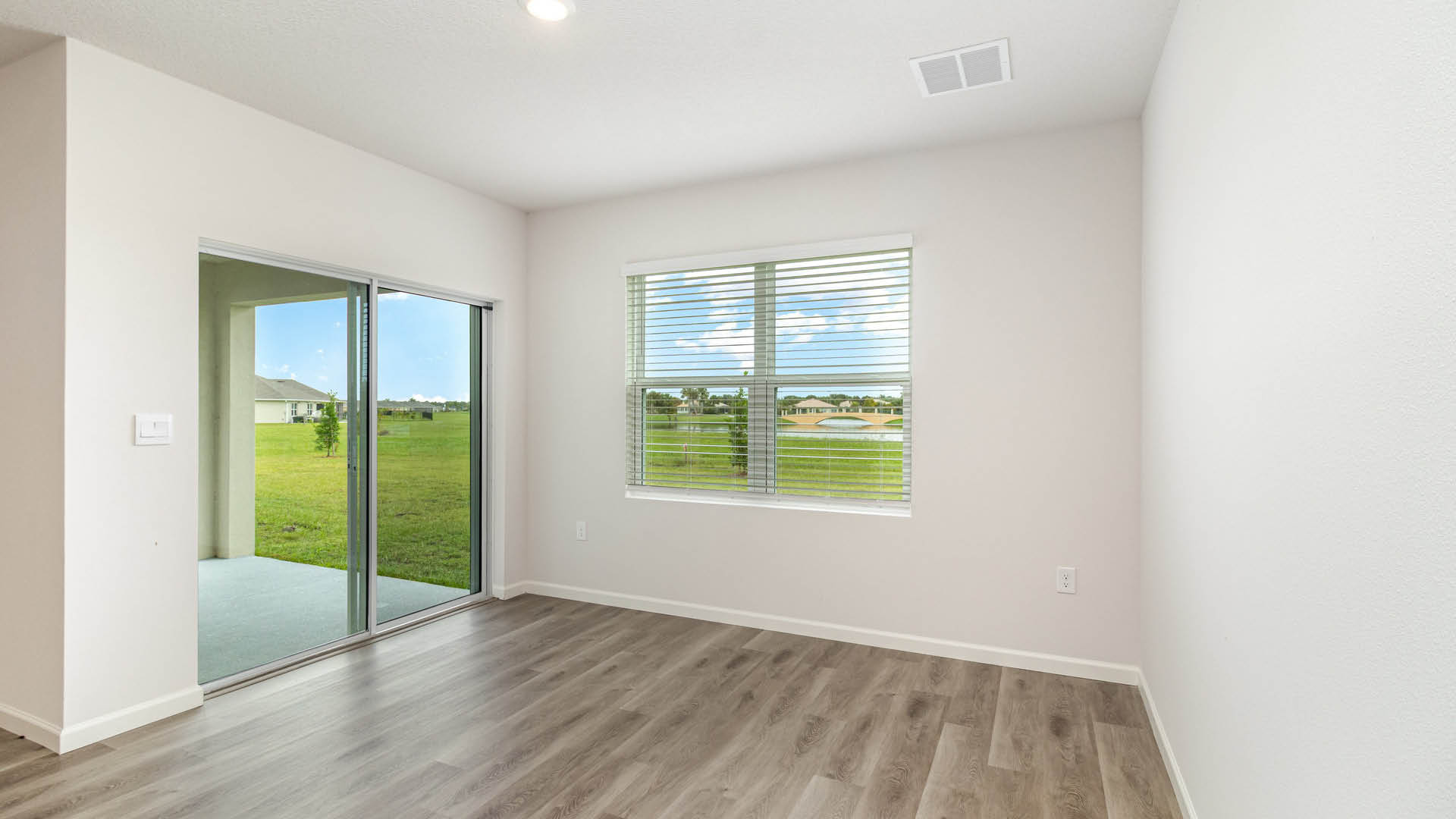 An empty room with white walls and LVP flooring features a window with blinds and glass sliding doors that look out onto a grassy lawn and patio area.