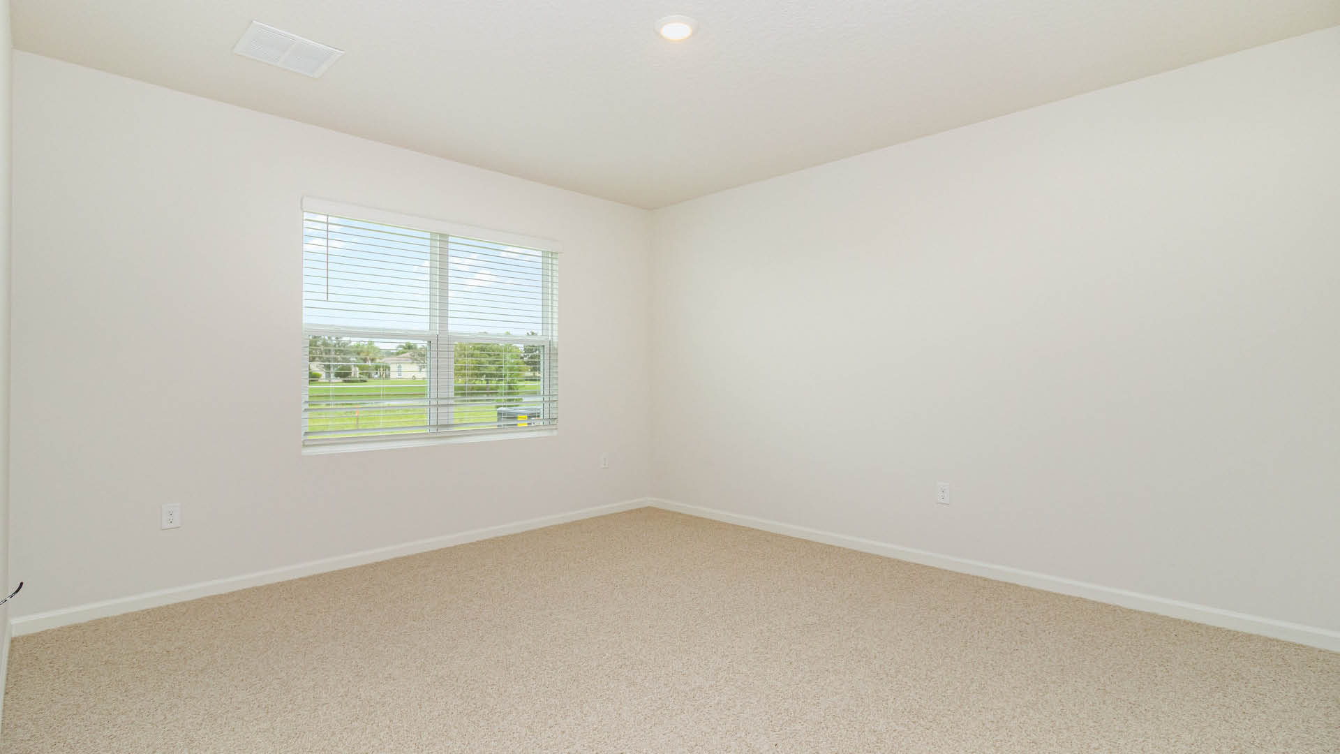 An empty room with white walls and beige carpeting features a single recessed ceiling light and a window with blinds overlooking a green lawn.
