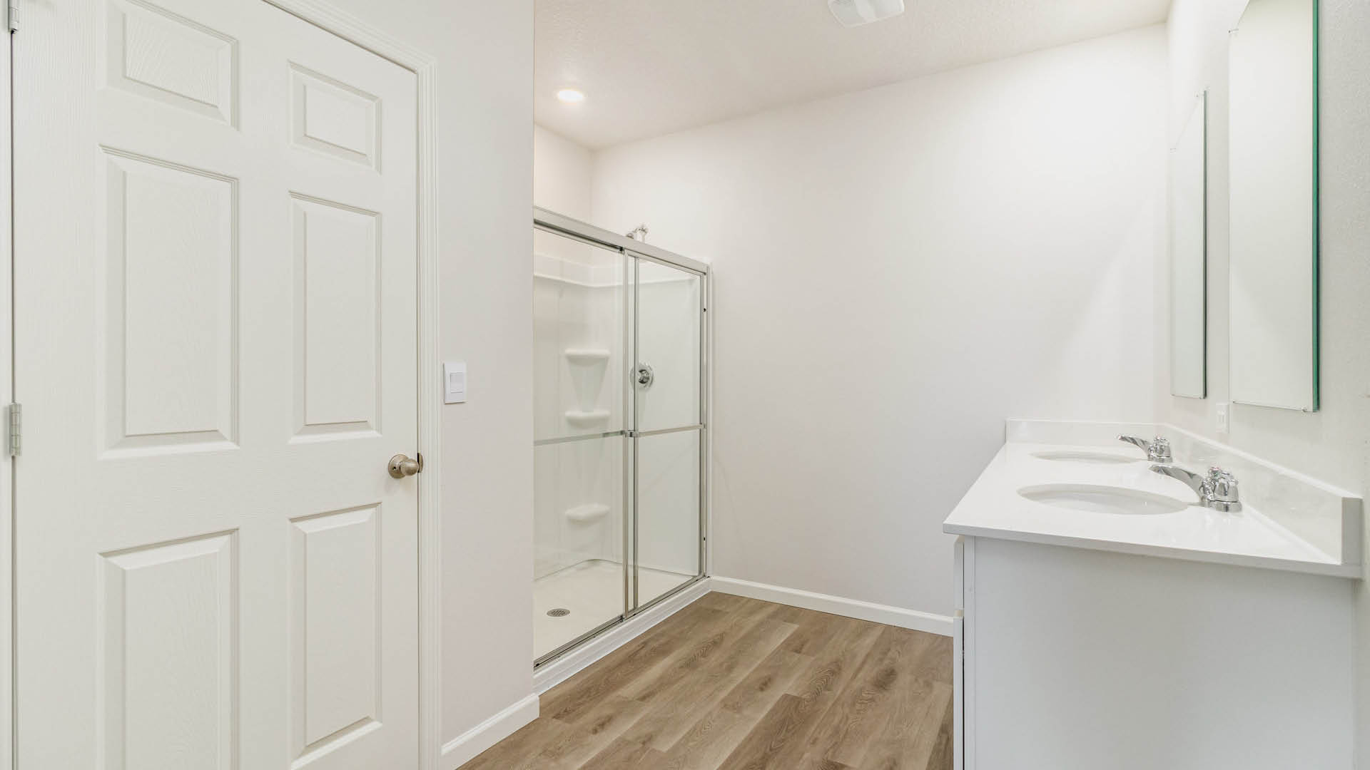 A white-walled bathroom with light brown LVP flooring features a double-sink vanity with large mirrors on the right, a glass-door walk-in shower in the background, and a closed white interior door on the left.