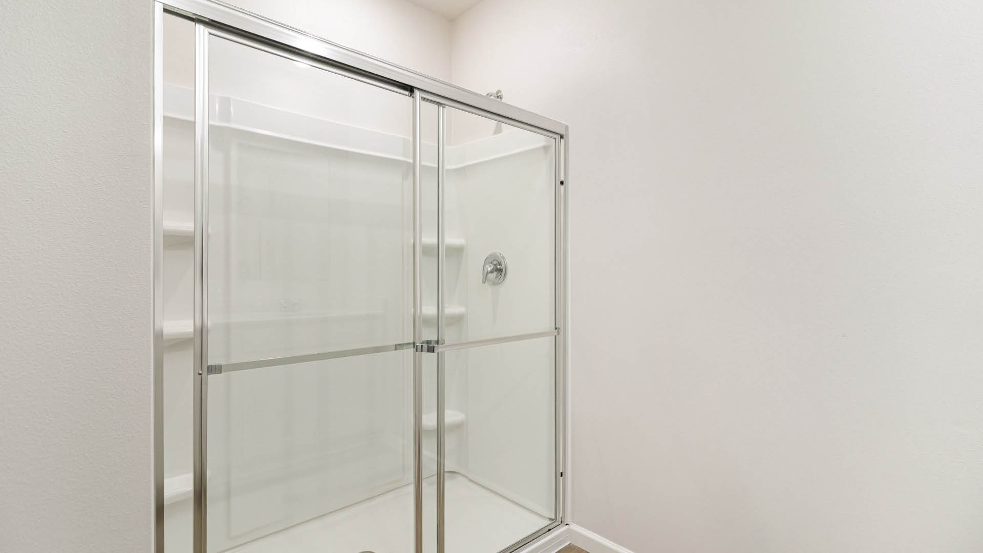 A close-up shot of a white walk-in shower stall features sliding glass doors with a silver metallic frame against a plain white wall.