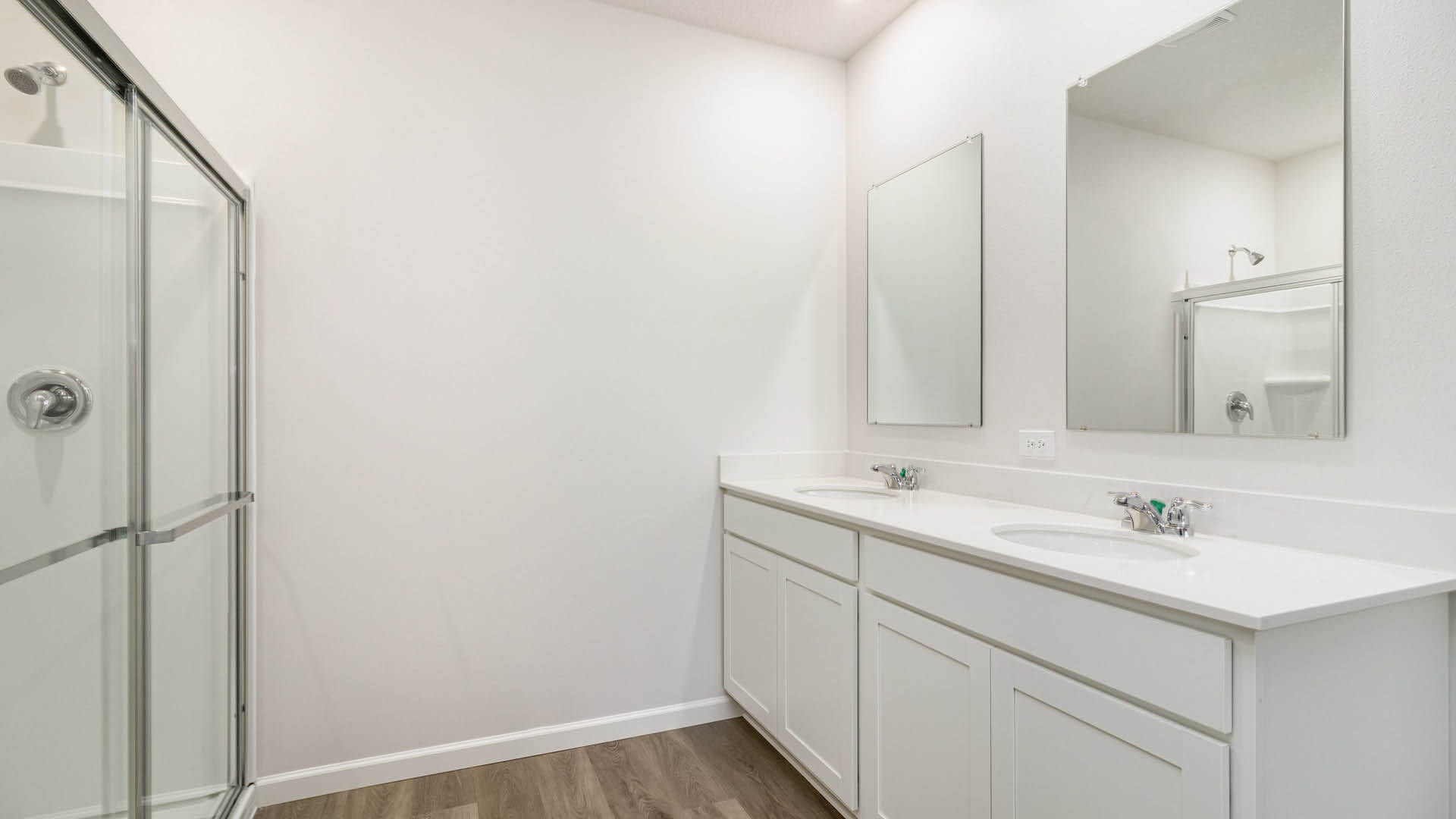 A white-walled bathroom with LVP flooring features a long white double-sink vanity with two mirrors on the right and a glass-enclosed walk-in shower on the left.