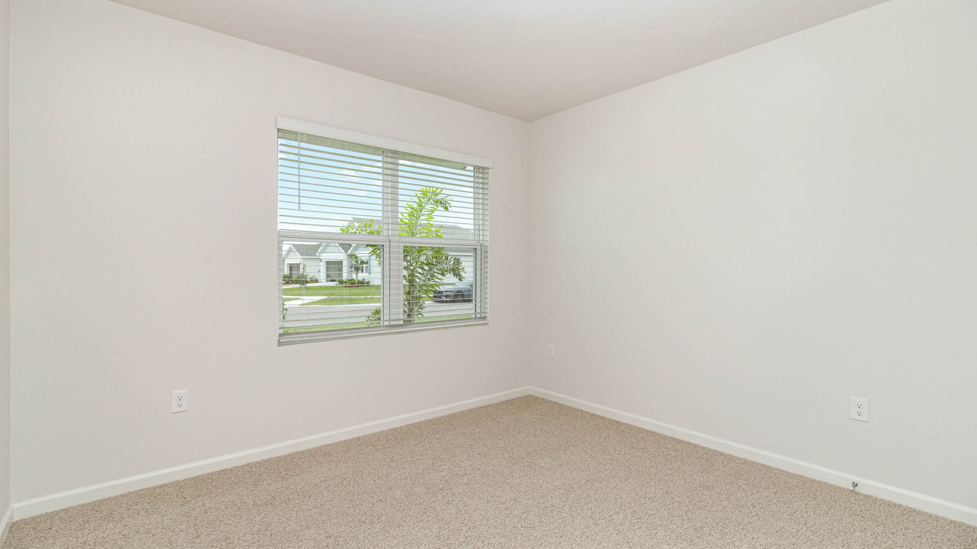 An empty room with white walls and beige carpeting features a single window with white blinds on the left wall.
