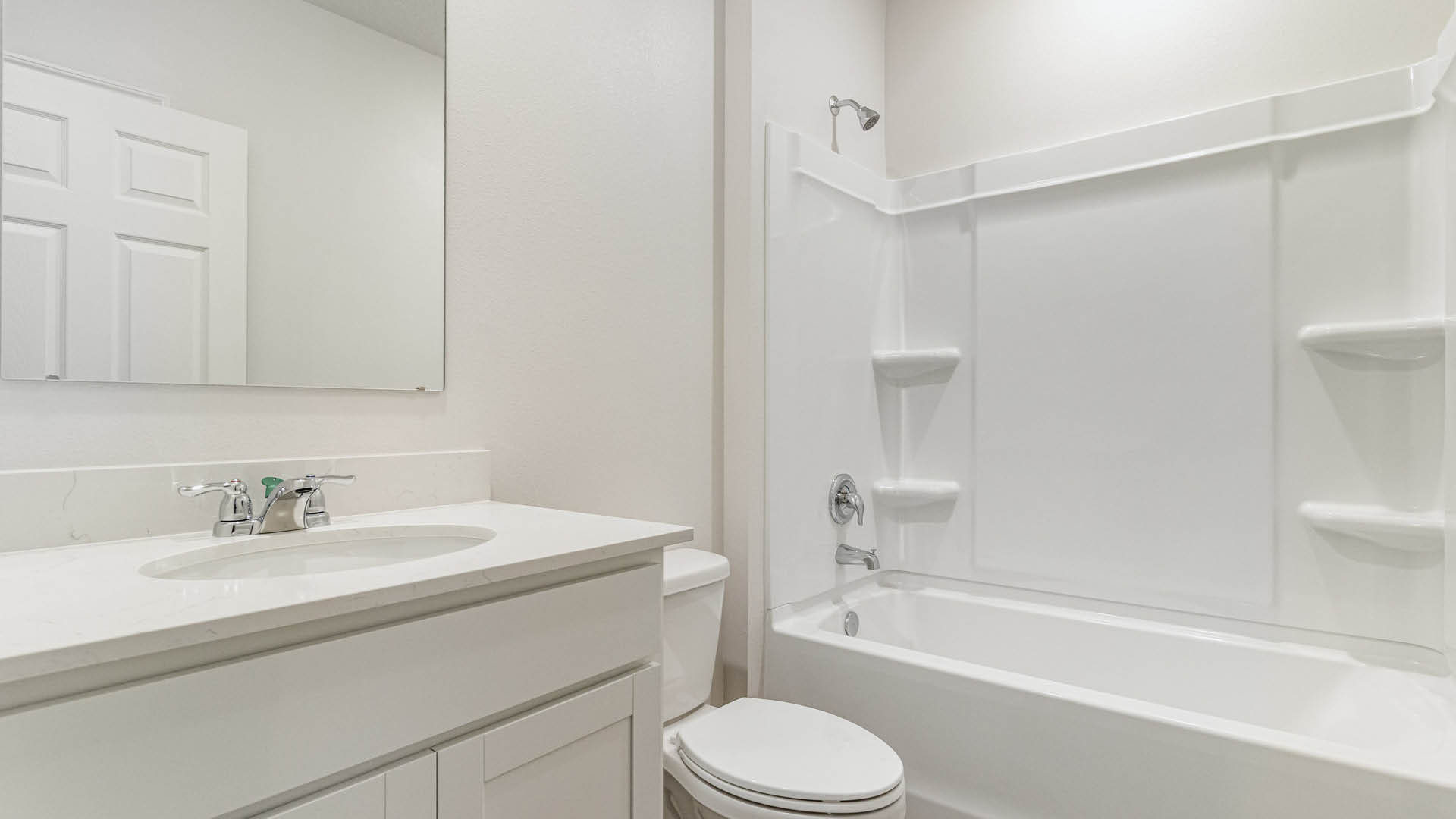 A white-walled bathroom features a single vanity with a chrome faucet and mirror on the left, a toilet in the center, and a combined white bathtub and shower unit on the right.