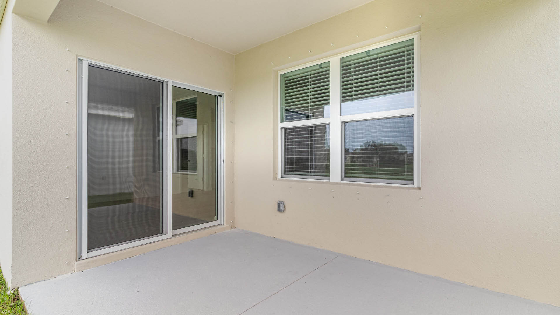 A covered concrete patio with beige walls features sliding glass doors with a screen on the left and a double-pane window with white blinds on the right.