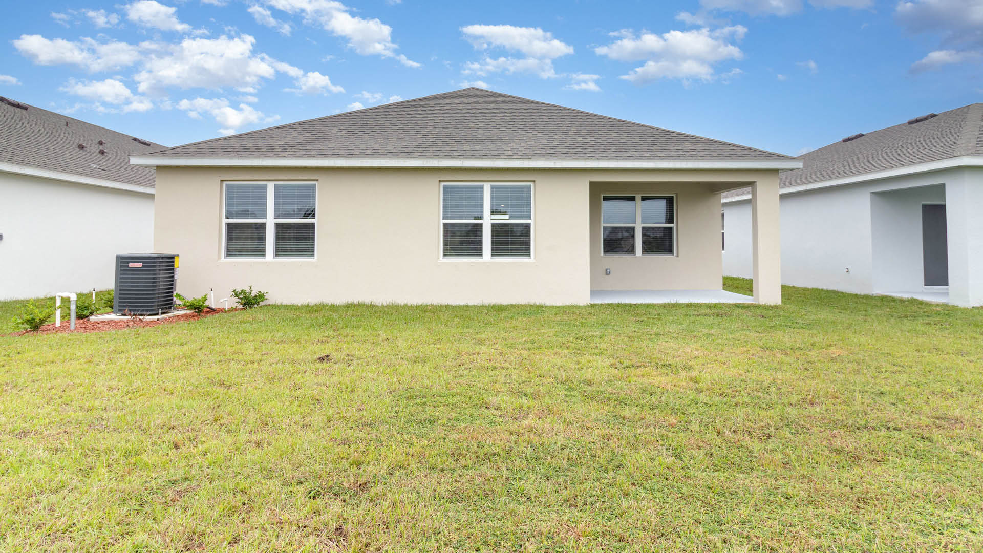 The back exterior of a single-story beige house features a grassy backyard, two large double windows, a small covered patio, and an outdoor air conditioning unit under a blue sky with scattered clouds.