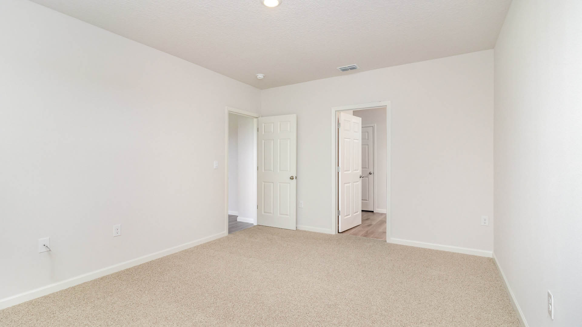 9. A spacious, empty room with white walls and beige carpet, featuring a closed door and a hallway leading to another area.