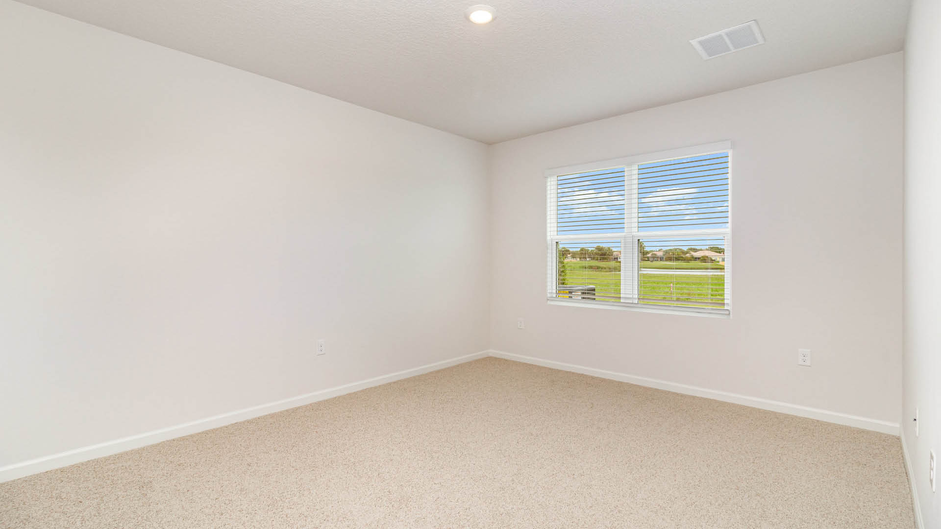Bright, empty room with beige carpet and white walls, featuring a window with blinds and natural light streaming in.