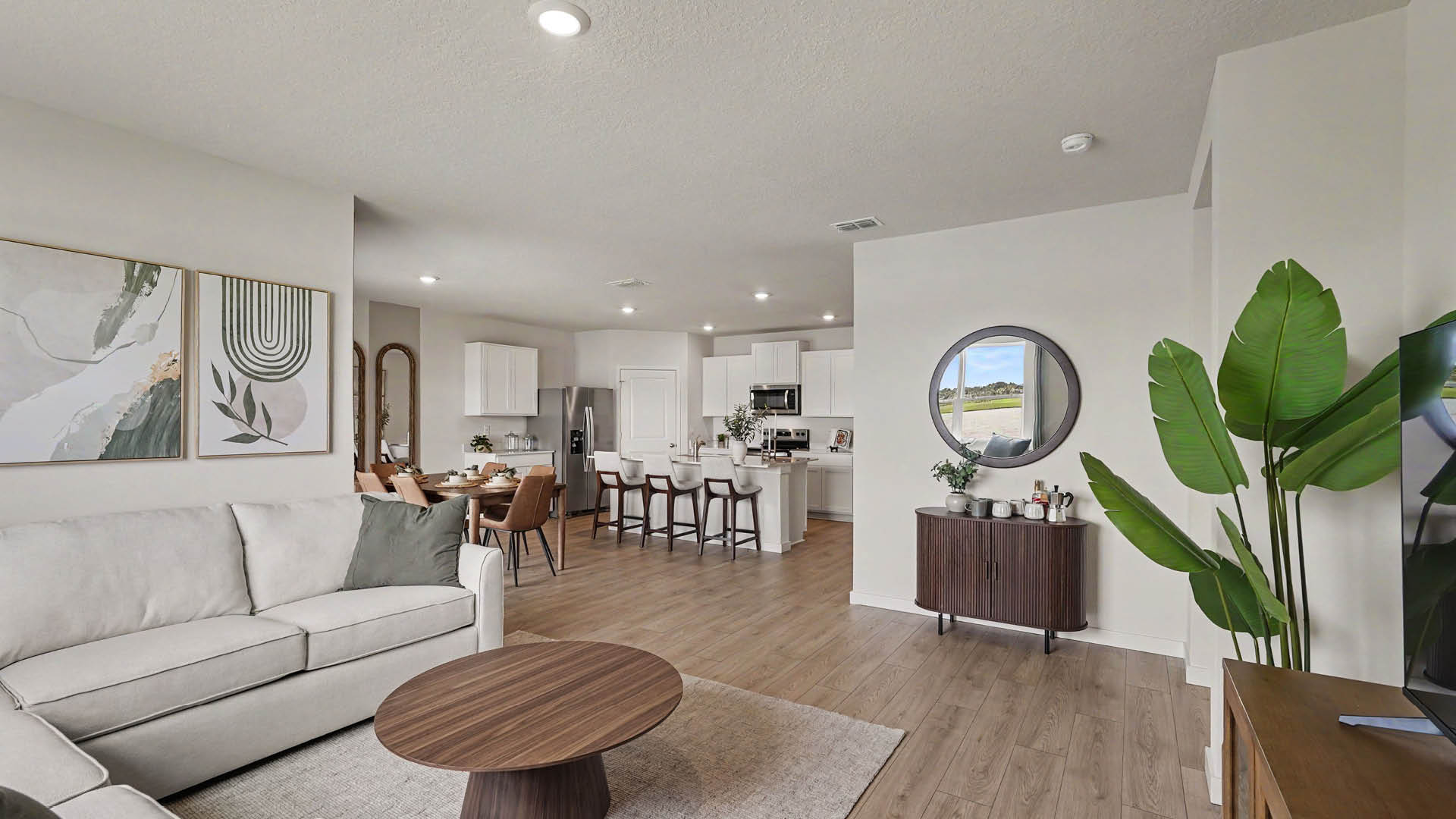 A modern open-concept living room and kitchen area features a light gray sofa, round wooden coffee table, dining table with chairs, bar stools at a kitchen island, and a large green plant next to a TV.