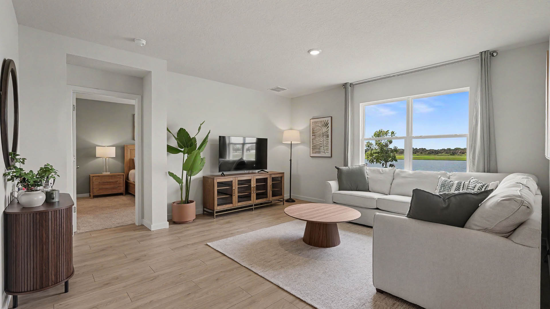 A modern living room features a light-colored sectional sofa with cushions, a round wooden coffee table on a rug, a TV on a wooden stand, a tall floor lamp, and a large window with a view of trees and water.