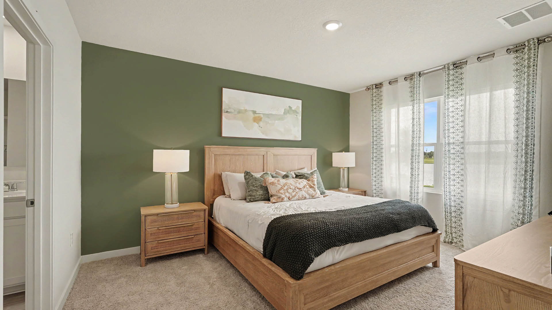 _ A neatly made bed with wooden frame and nightstands is set against a green accent wall with a landscape painting above, flanked by two lamps and sheer curtains covering a window.