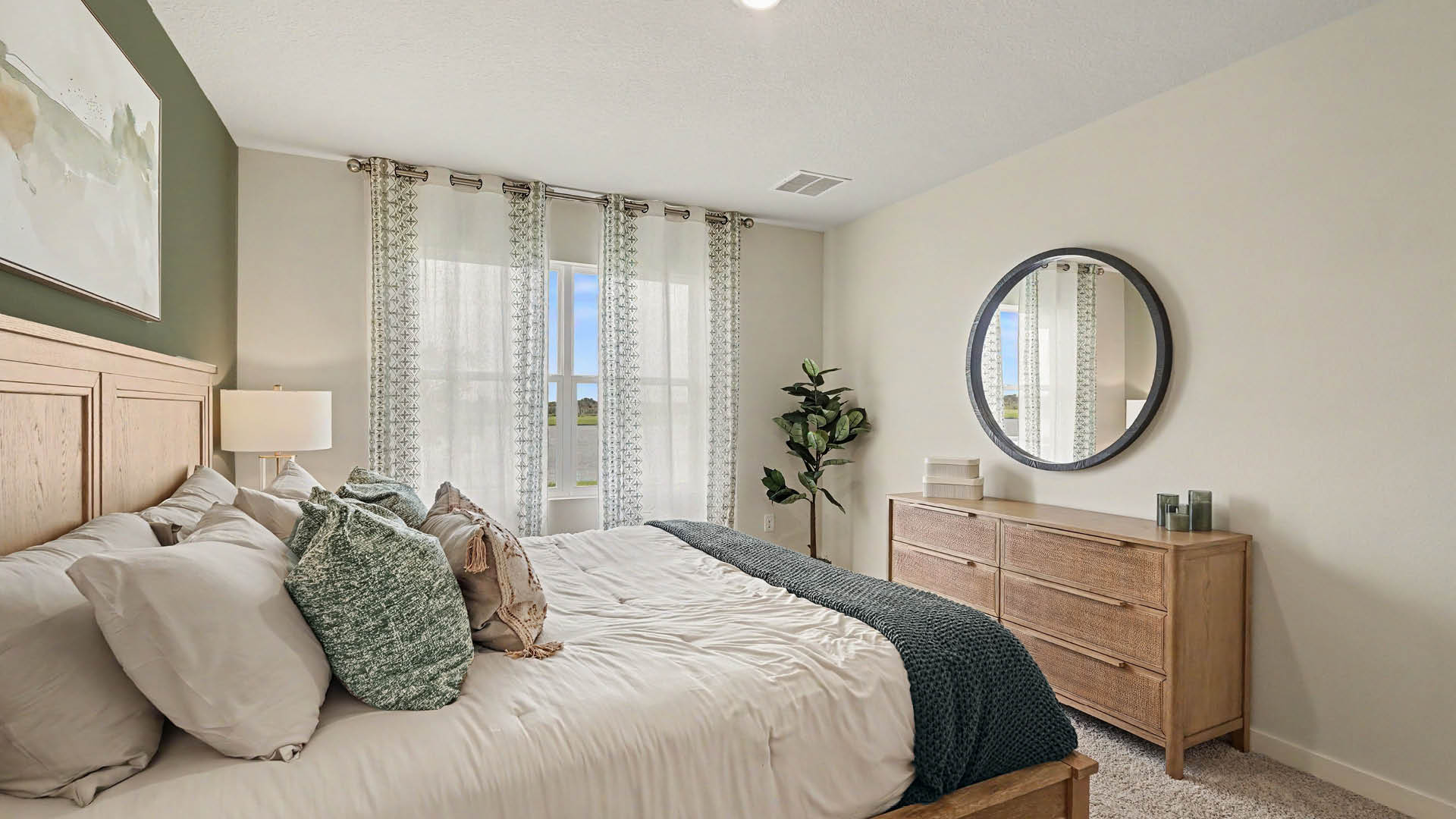 A neatly made bed with multiple pillows and a dark throw blanket is positioned next to a window with sheer curtains, opposite a wooden dresser with a round mirror and decorative items in a softly lit bedroom.