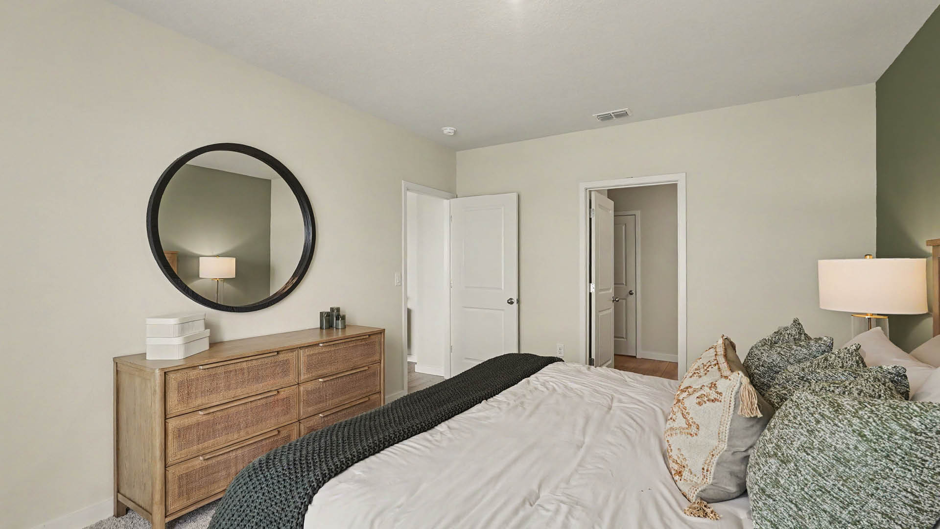 A neatly made bed with multiple pillows faces a wooden dresser topped with decorative items and a round mirror in a softly lit bedroom.
