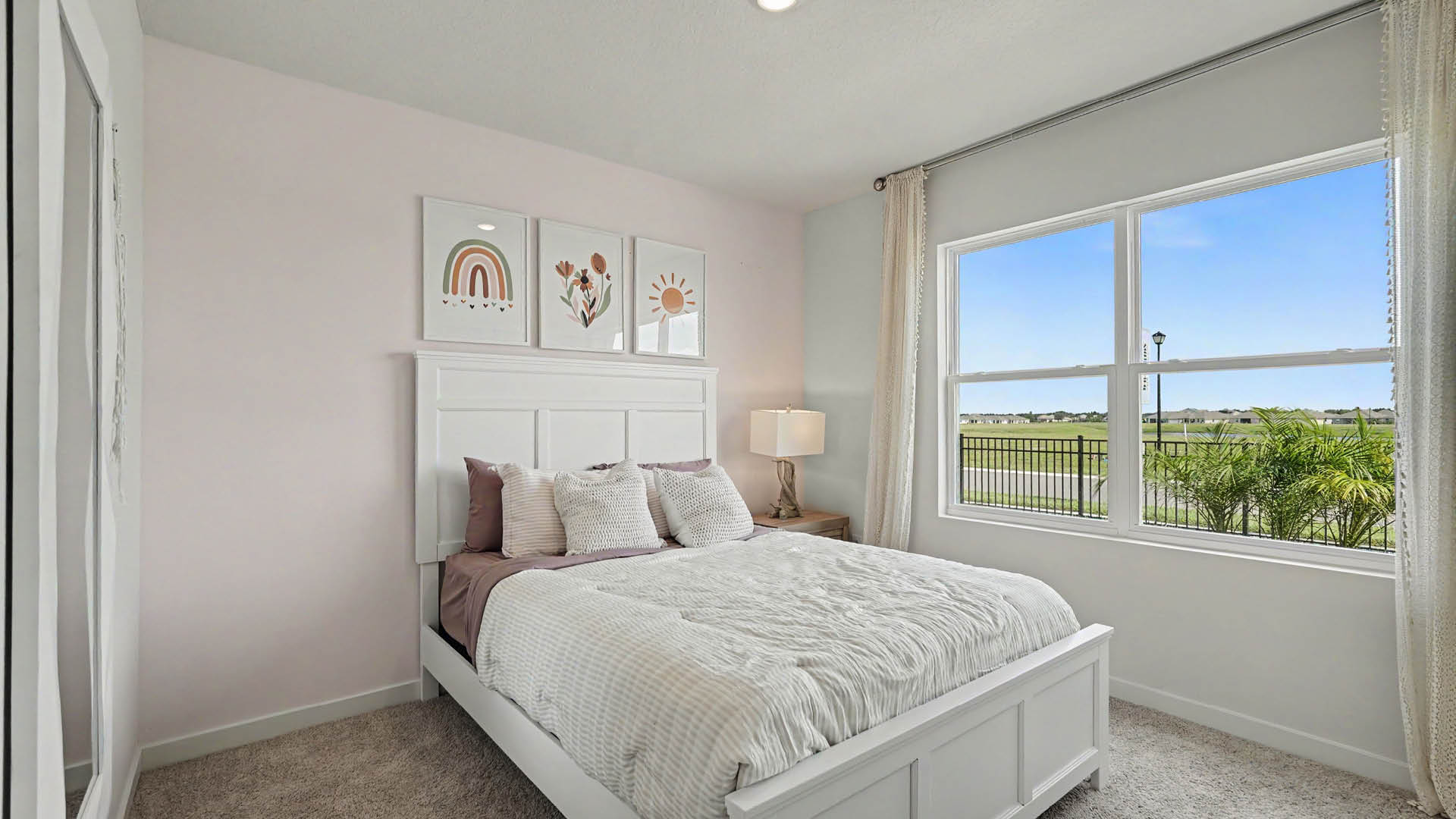 A neatly made bed with white and mauve bedding is positioned against a light-colored wall with three framed artworks above it, next to a window showing a clear outdoor view and a bedside table with a lamp.