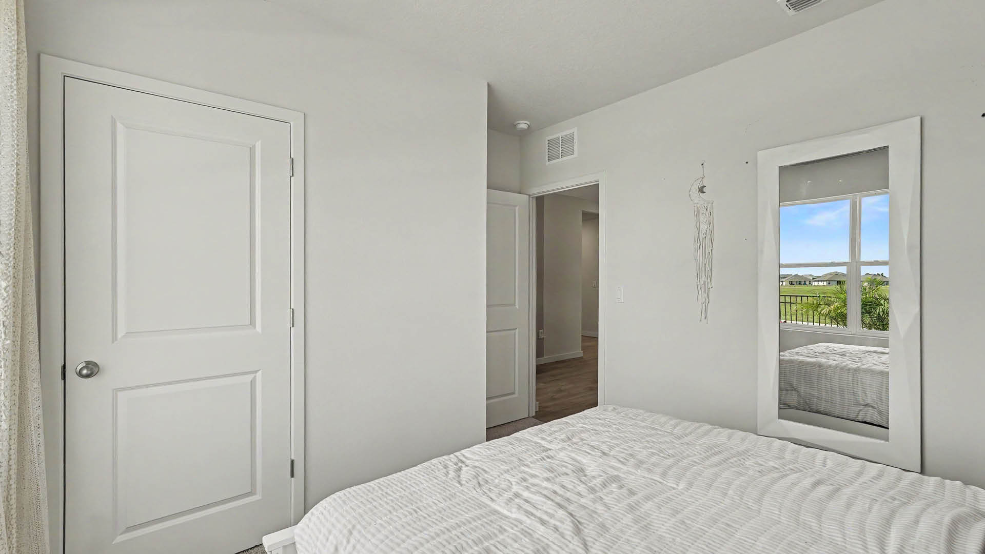 A minimalist bedroom features a neatly made bed with white bedding, a closed white door, an open doorway leading to another room, and a wall-mounted mirror reflecting a window with a view of greenery outside.