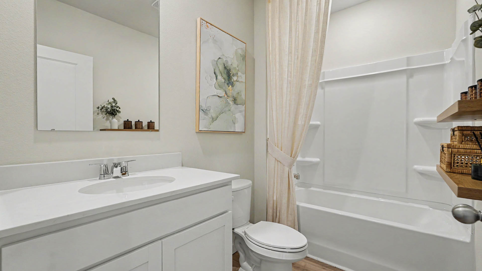 A clean, modern bathroom features a white vanity with a sink, a toilet, a bathtub with a shower curtain, a large mirror, a framed abstract painting, and wooden shelves holding wicker baskets.