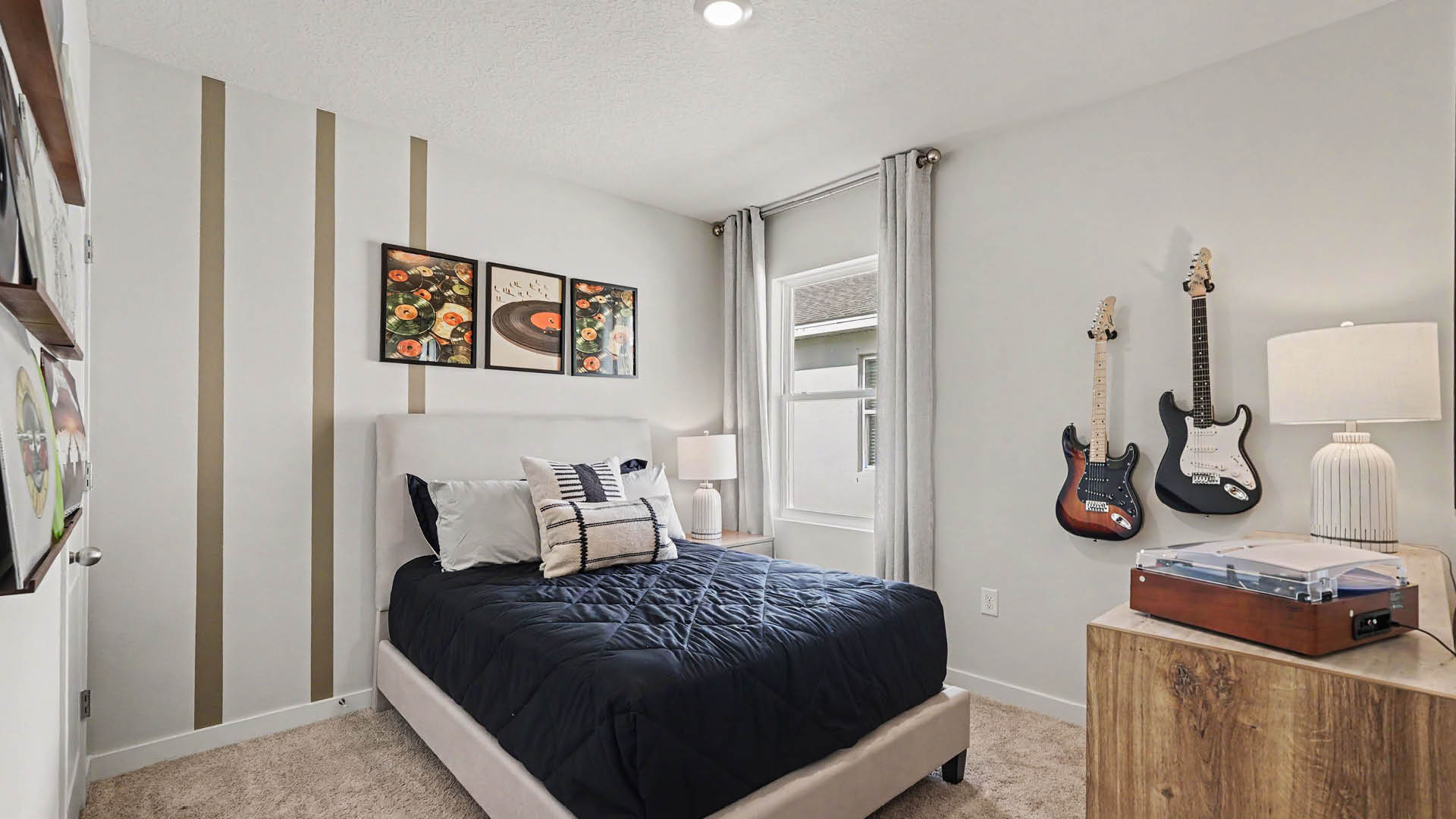 A modern bedroom features a bed with dark bedding and decorative pillows, two electric guitars mounted on the wall, a wooden dresser with a lamp and a record player, and a window with light curtains.