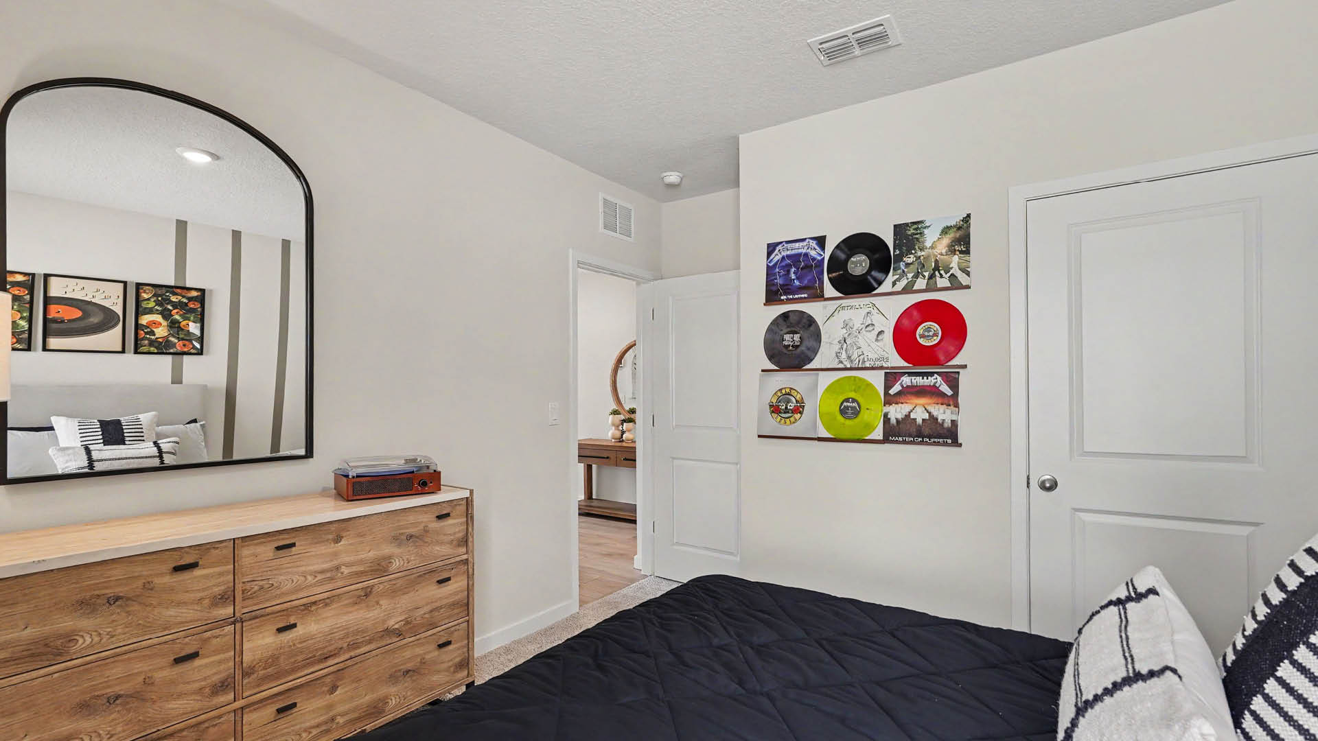 A bedroom features a bed with dark bedding and pillows, a wooden dresser with a mirror above it, and a wall decorated with various vinyl records.